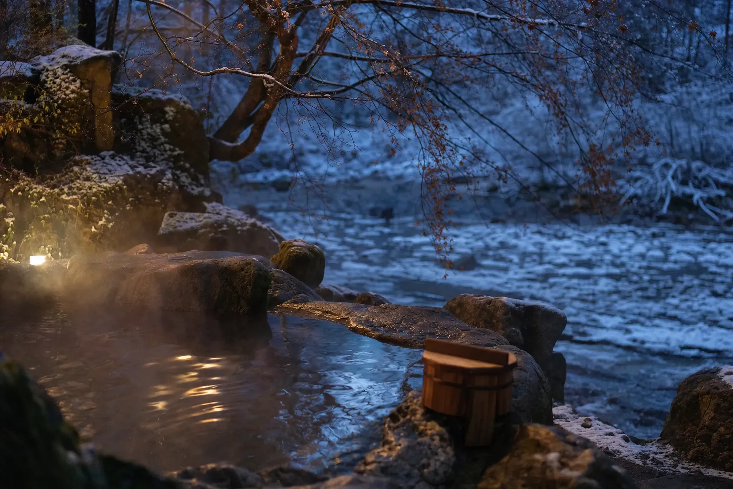 A riverside open-air rotenburo bath at a Kurokawa Onsen ryokan, mineral-tinged water against mossy stone and overhanging trees