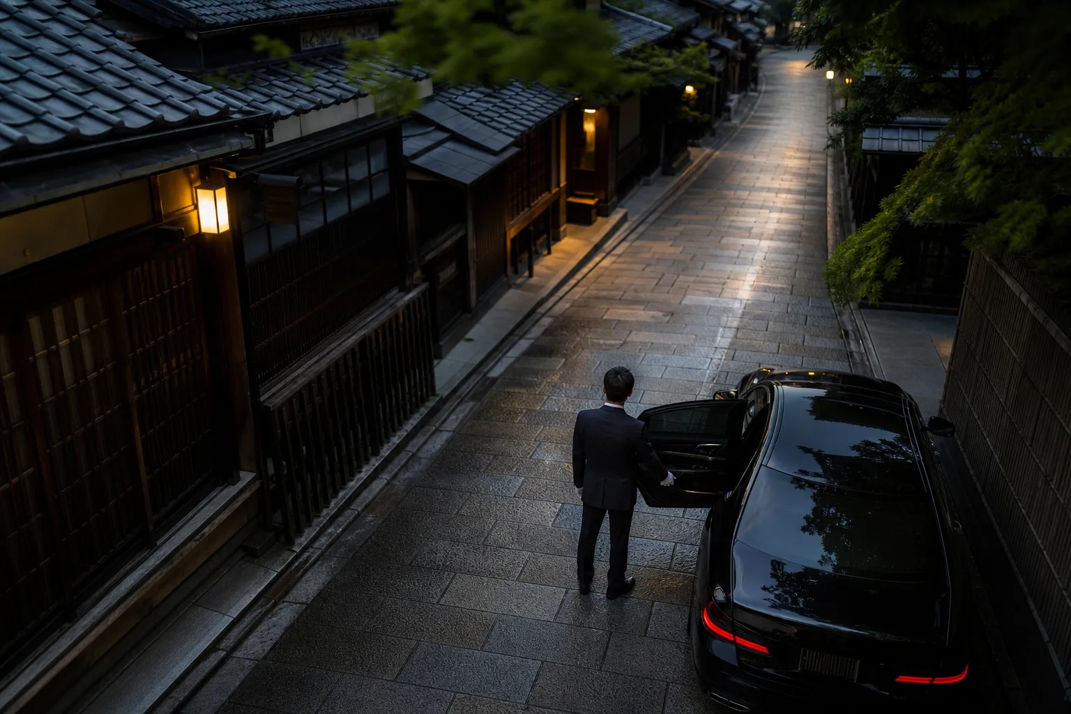 A man in a dark suit stands beside a black luxury car parked on a traditional Japanese street with wooden buildings.