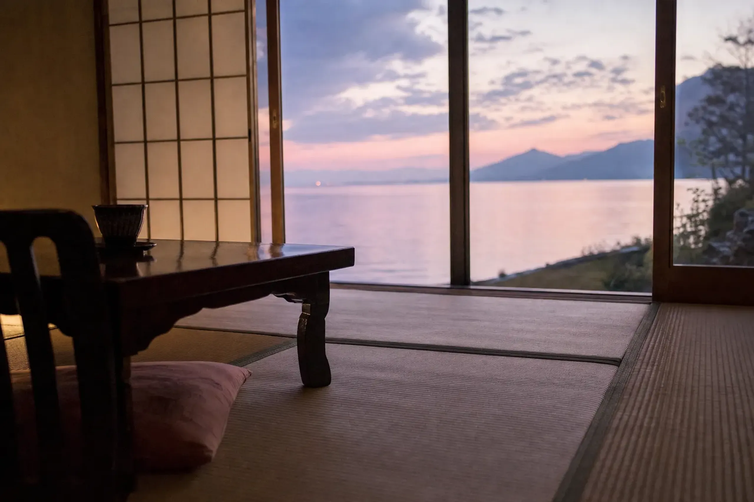 A lakeside ryokan room overlooking Lake Biwa at dusk, tatami floor and low table with the still water visible through the window
