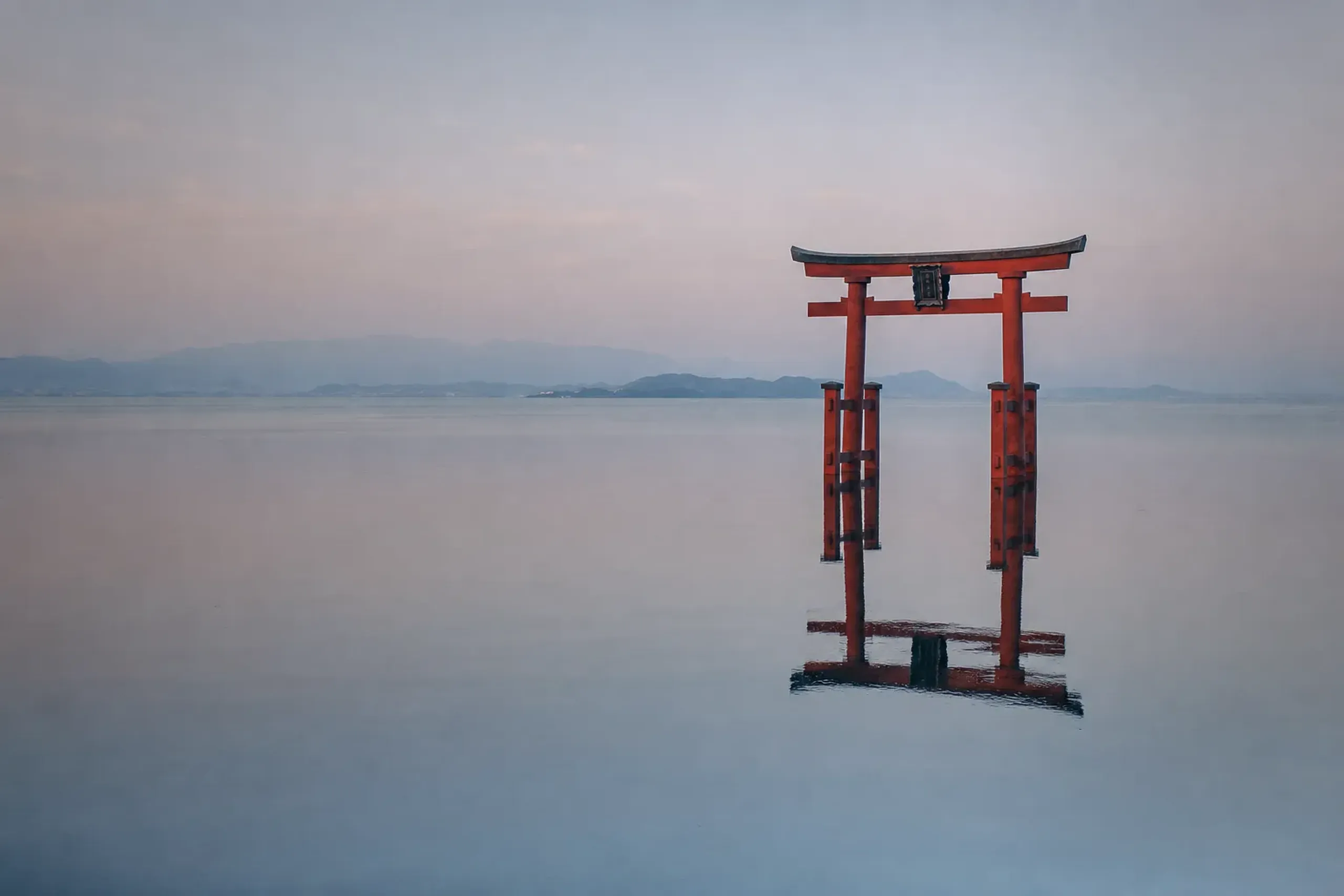 Shirahige Shrine's vermilion torii gate rising directly from the still waters of Lake Biwa at dusk, silhouetted against the hazy far shore