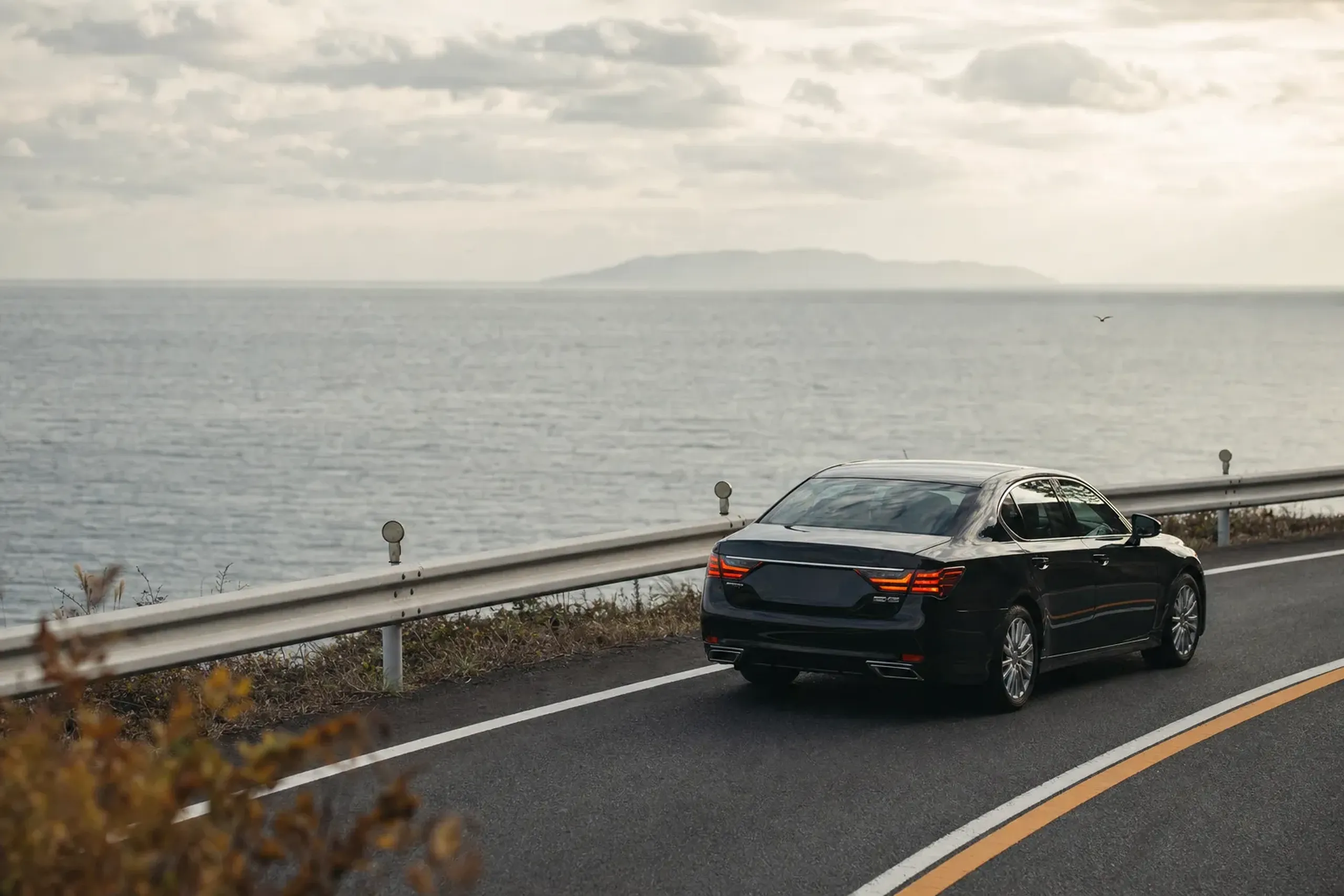 A black chauffeured sedan on the coastal road approaching Munakata City, the Genkai Sea visible beyond