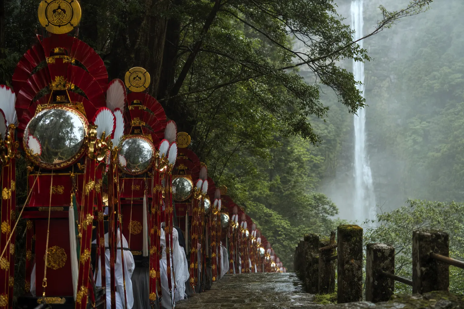 The twelve tall, narrow ogi mikoshi fan shrines decorated with fans and mirrors processing along the stone-step approach at Kumano Nachi Taisha Grand Shrine, with Nachi Waterfall cascading in the back