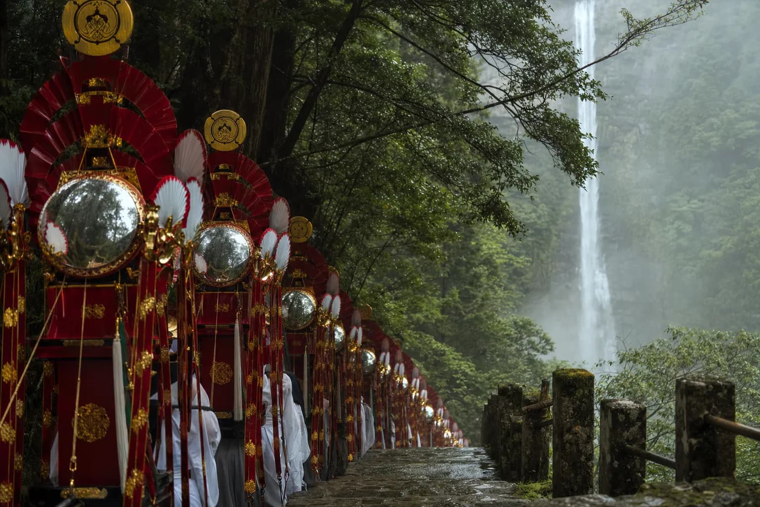The twelve tall, narrow ogi mikoshi fan shrines decorated with fans and mirrors processing along the stone-step approach at Kumano Nachi Taisha Grand Shrine, with Nachi Waterfall cascading in the back