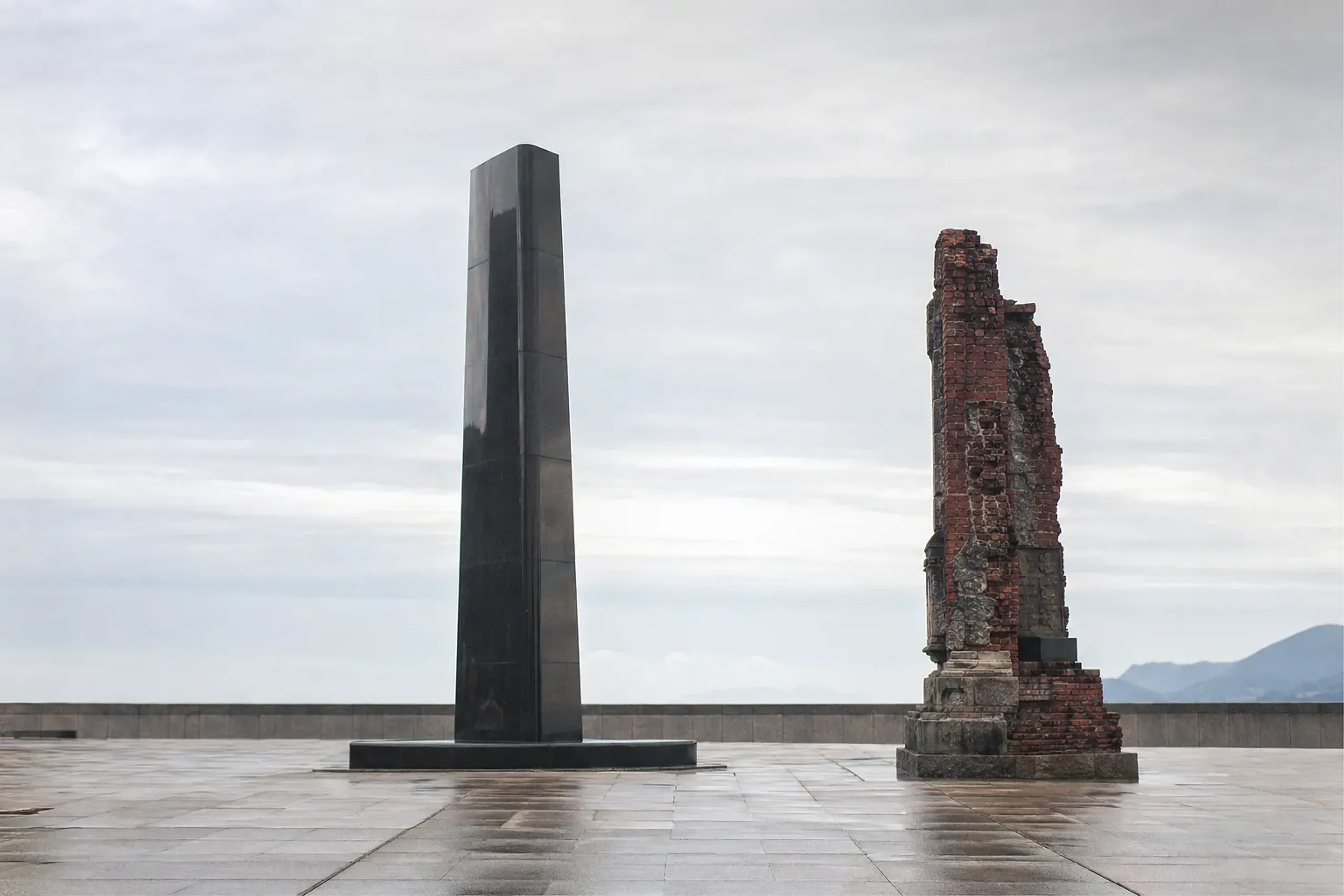 The black stone monolith marking the atomic bomb epicenter at Hypocenter Park in Nagasaki, with the preserved damaged brick pillar from the former Urakami Cathedral standing beside it