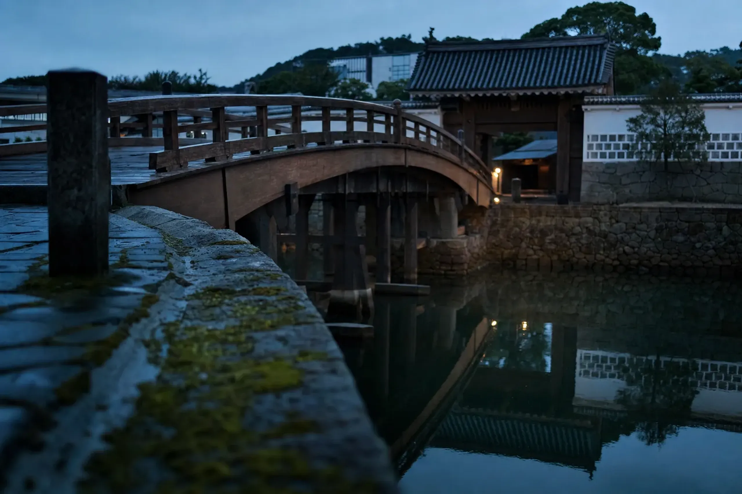 the arched Omotemon Bridge crossing into Dejima's eastern entrance, framed by muted timber facades in the register of two hundred years ago