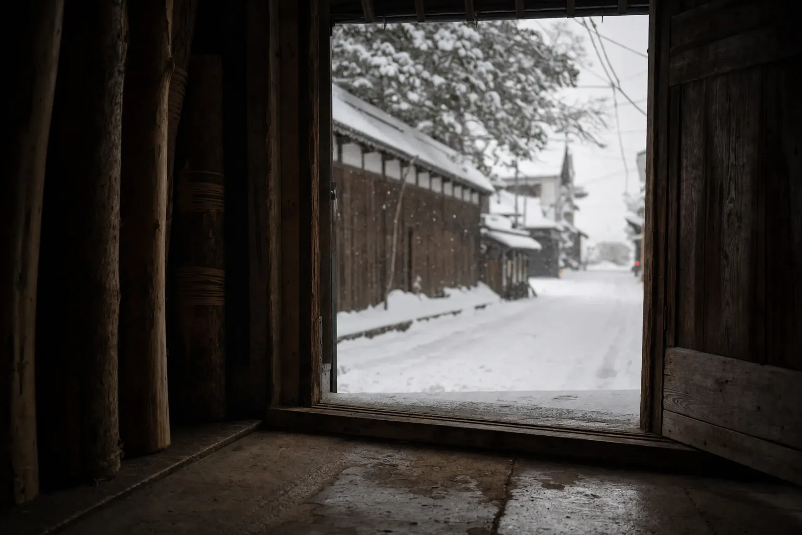 a sliding wooden brewery door standing open onto a snow-covered street in Niigata City near the Sea of Japan coast, cedar trunks with snow piled against them