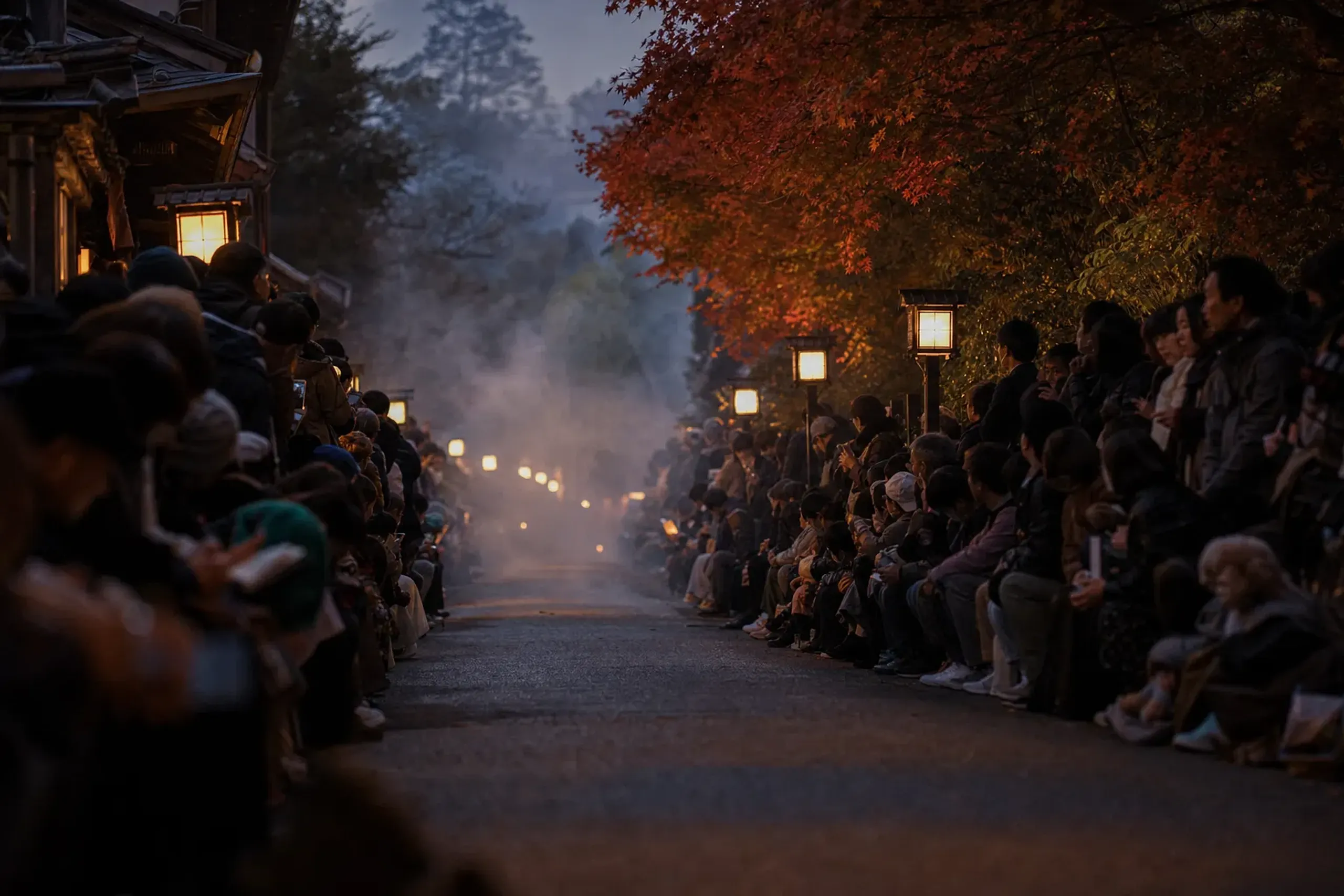 Dense crowds lining the narrow asphalt of Nishimonai Honcho Street at dusk with low-drifting bonfire smoke