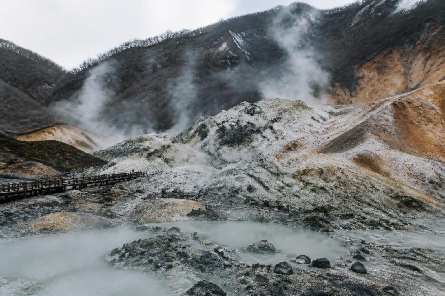 Wooden boardwalk winds through a steaming geothermal landscape with mineral-colored rocks and mist rising from hot pools at the base of barren hills.