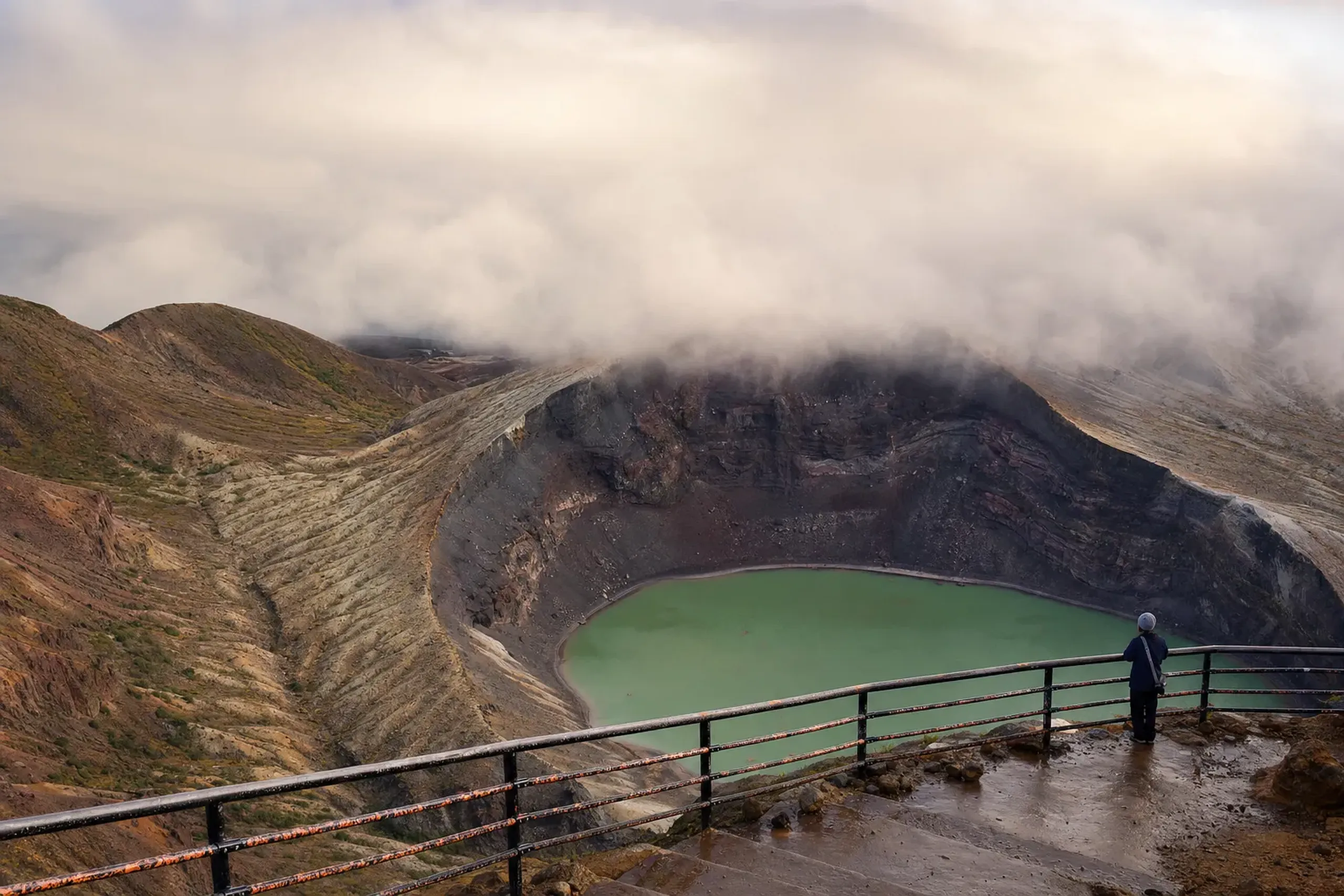 the railed observation deck overlooking Okama crater lake with thin cloud beginning to build over the emerald water