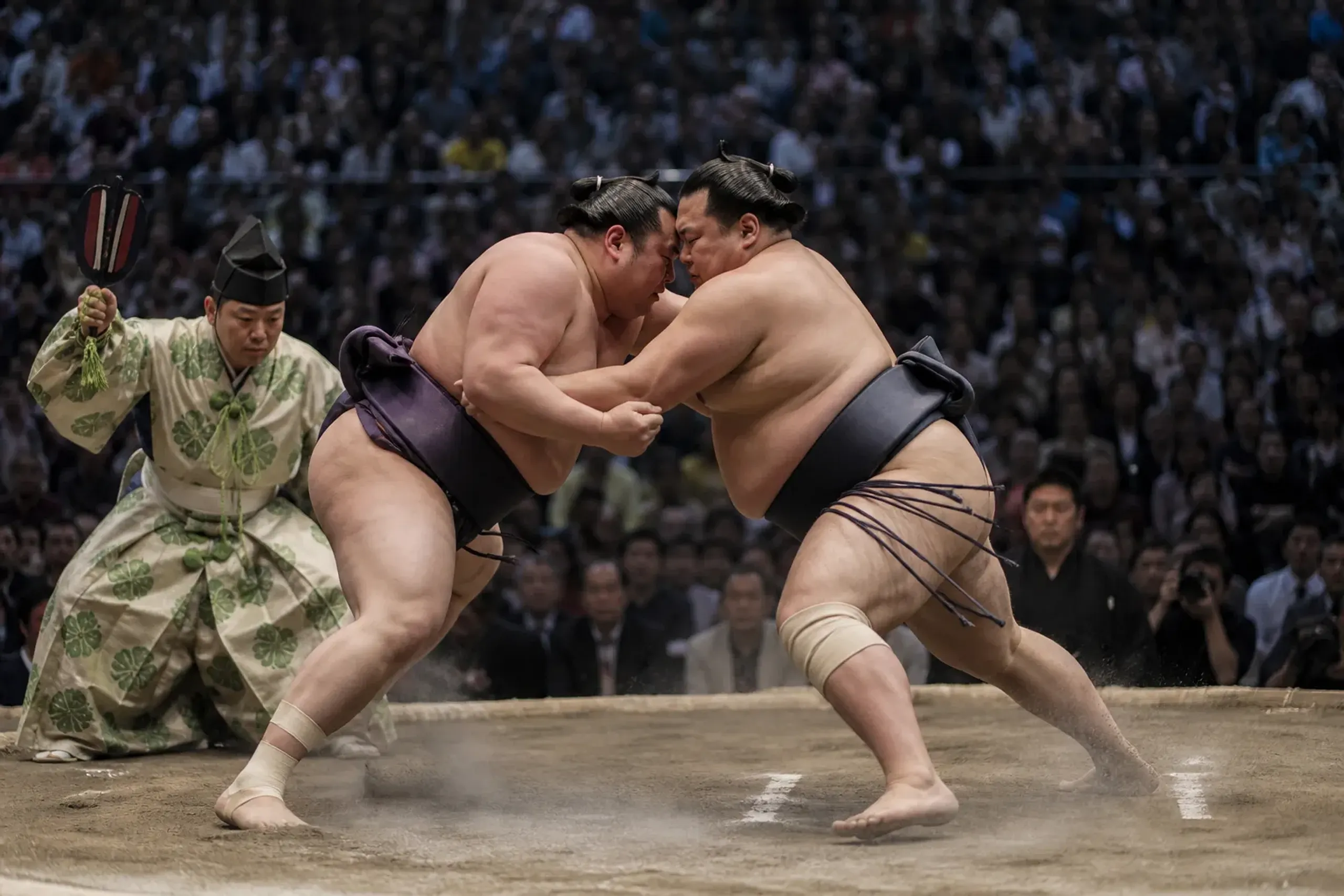 Two sumo wrestlers colliding on the clay ring during a top-division bout with a referee alongside