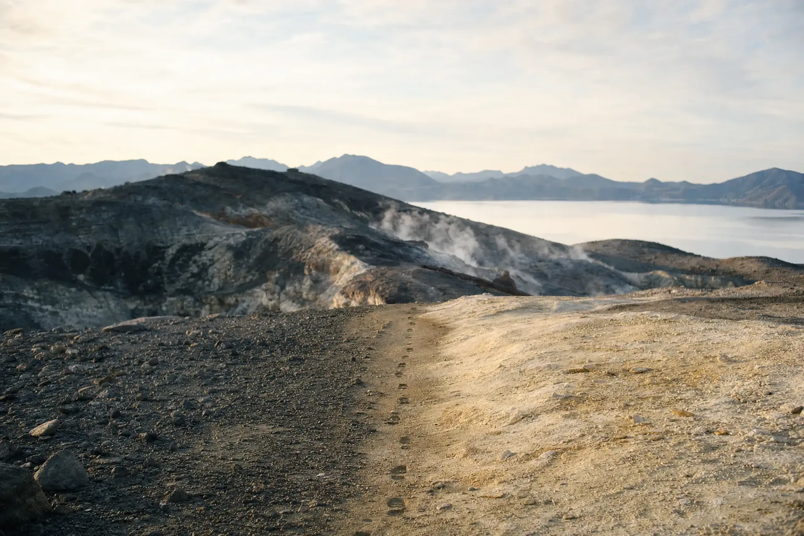 A walking path at Osorezan crossing from gray volcanic ash to warm pale stone, with steam vents rising and the white caldera lake visible ahead against dark rock