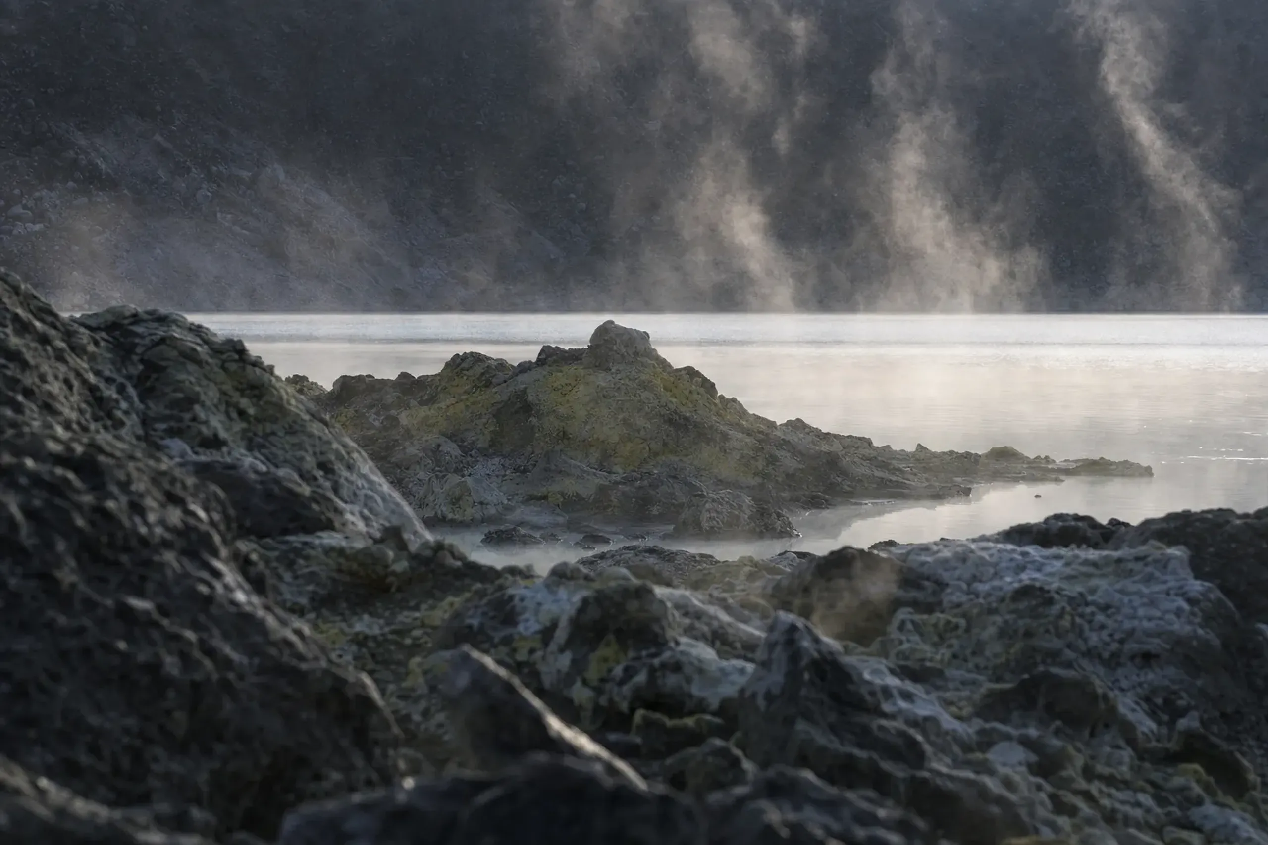 Autumn light low over the pale caldera lake of Osorezan, steam drifting visibly against the cool air with dark volcanic rock in the foreground