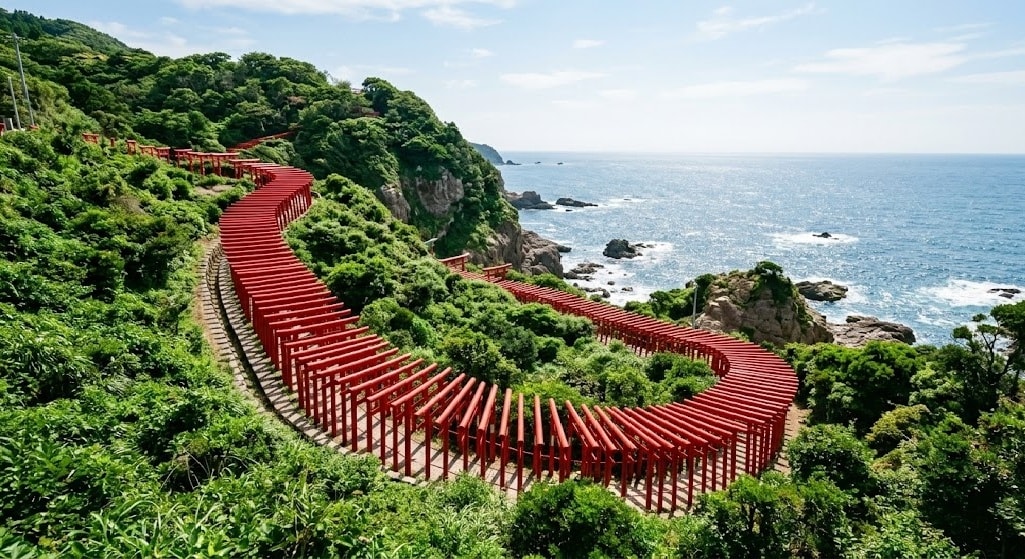 A line of bright red torii gates winding down a lush green cliff toward the blue ocean at Motonosumi Shrine.