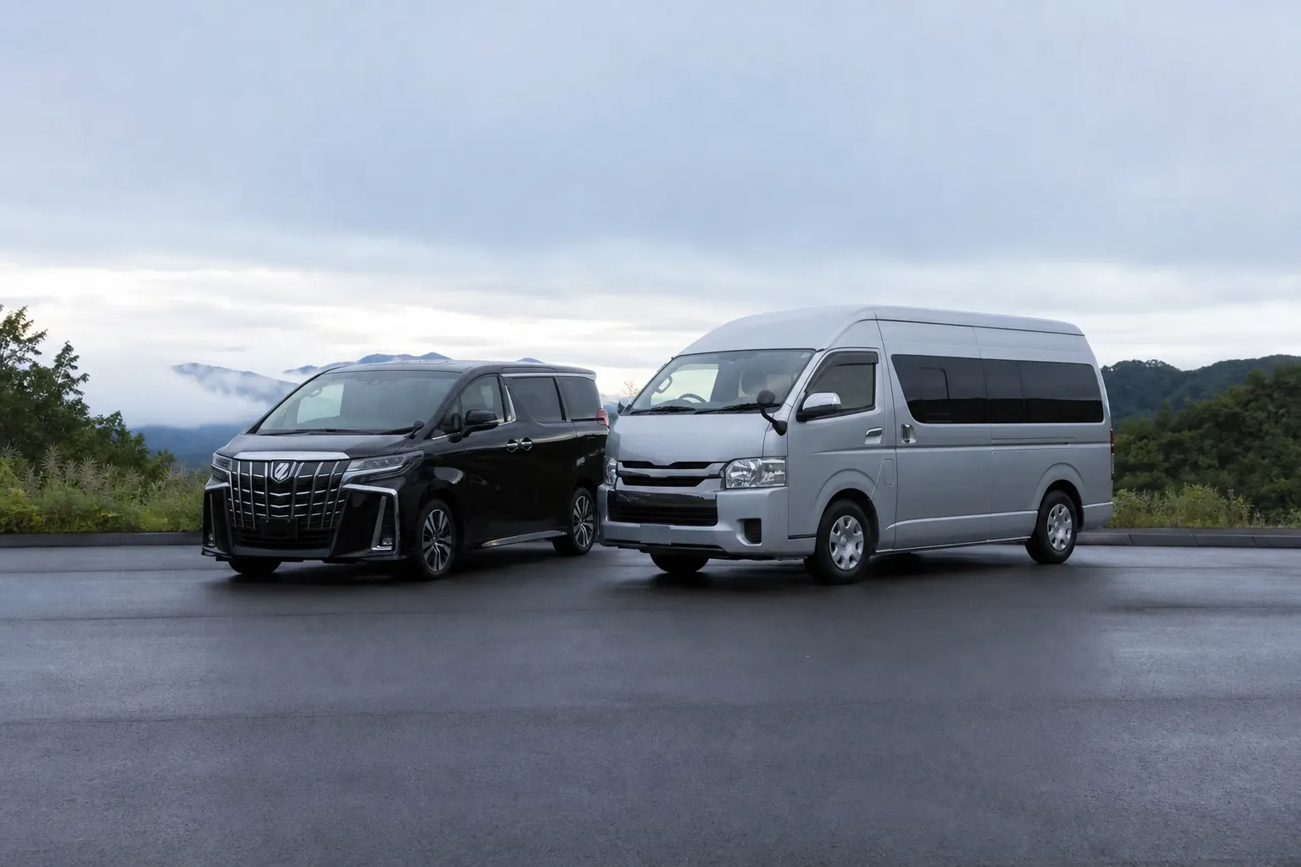 A black Toyota Alphard and a silver Toyota Hiace parked side by side on a clean asphalt lot in Japan