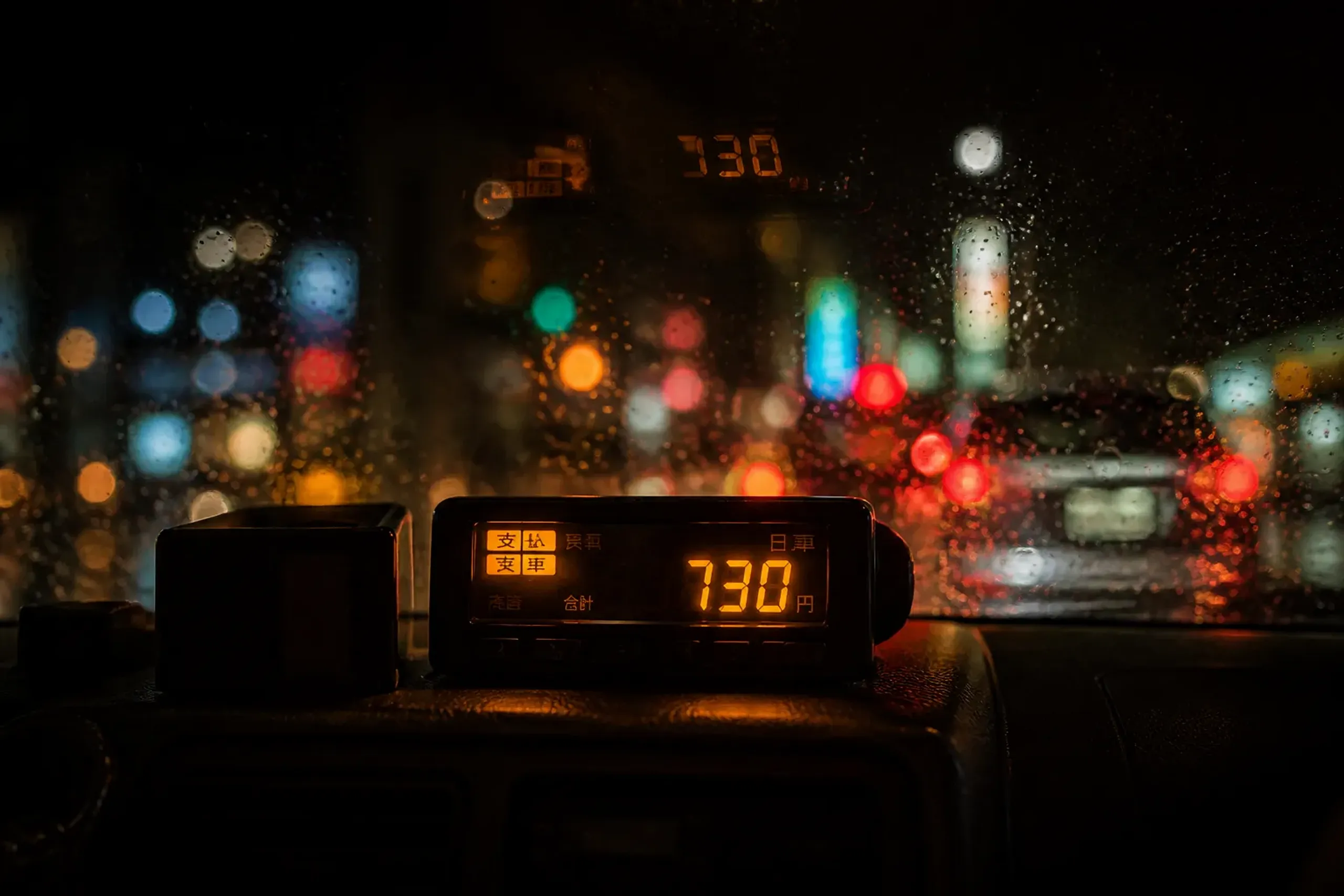 the taximeter mounted on the dashboard of a Japanese hire car, glowing with fare digits at night through the windshield