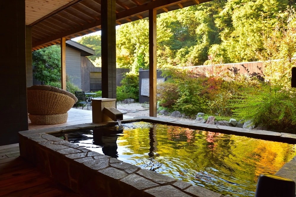 Stone hot tub on a wooden deck under a covered patio, surrounded by trees and a lush garden.