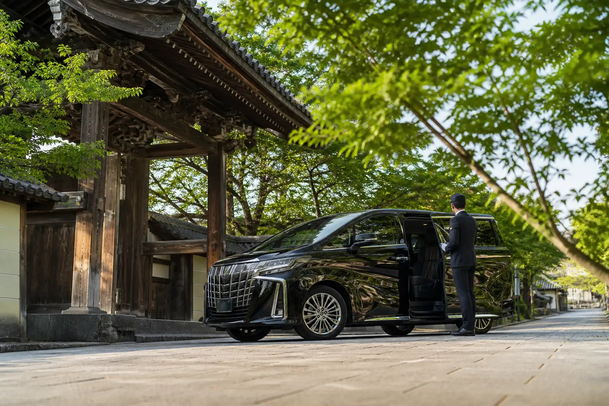 a black Toyota Executive Alphard parked outside a traditional wooden temple gate in Kyoto's Higashiyama district, its uniformed chauffeur standing beside the open rear door in early morning light