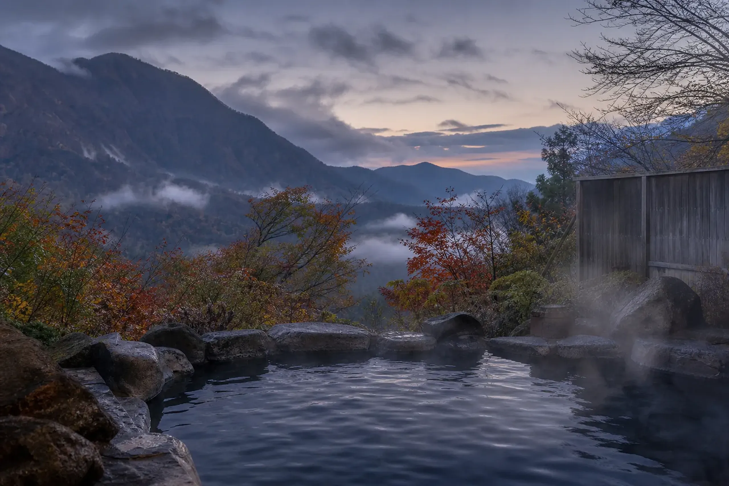 Steaming outdoor onsen bath surrounded by autumn mountain scenery at dusk