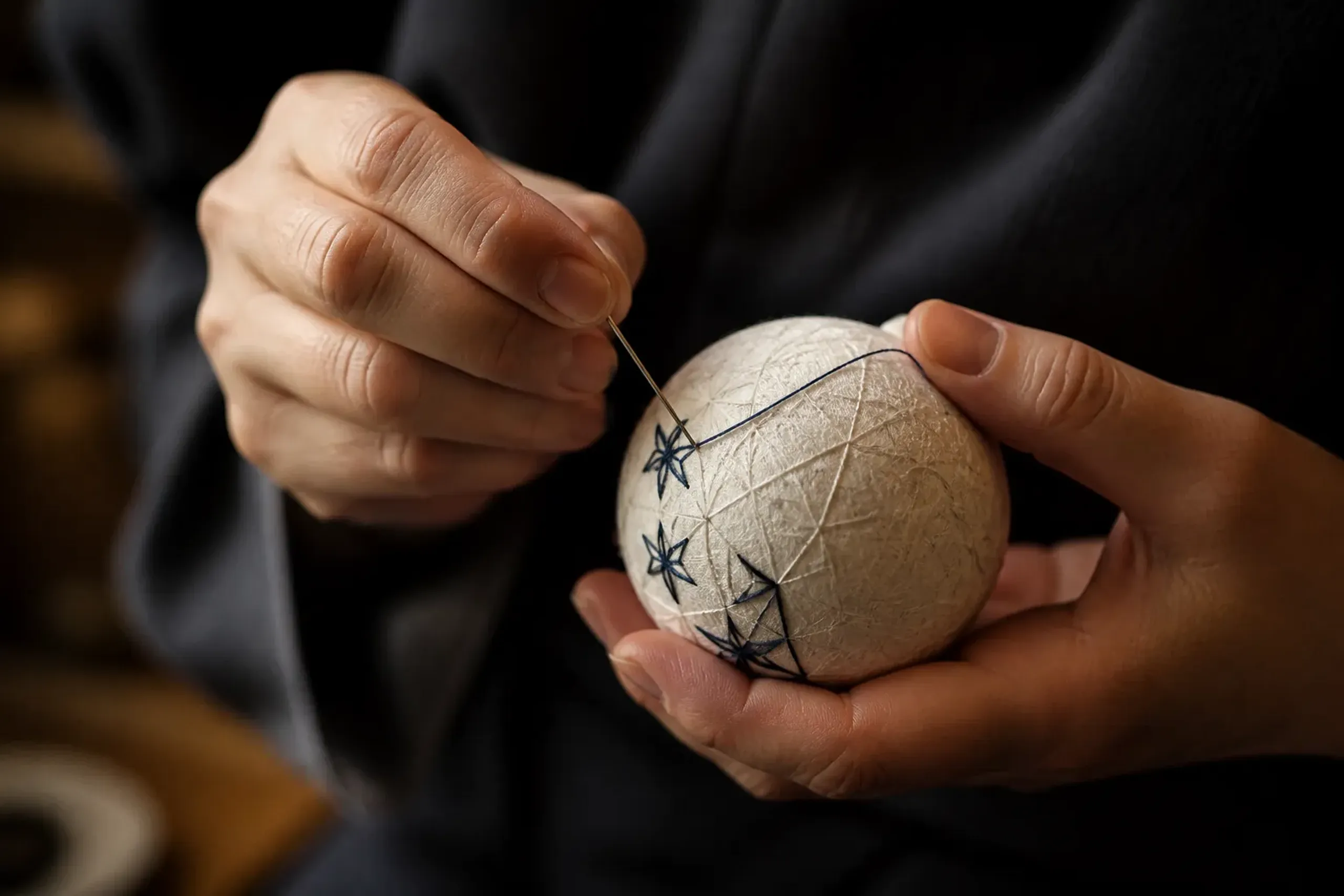 a wound cotton temari core held in hand with a threaded needle mid-stitch, the sphere marked into geometric divisions for the asanoha hemp-leaf pattern