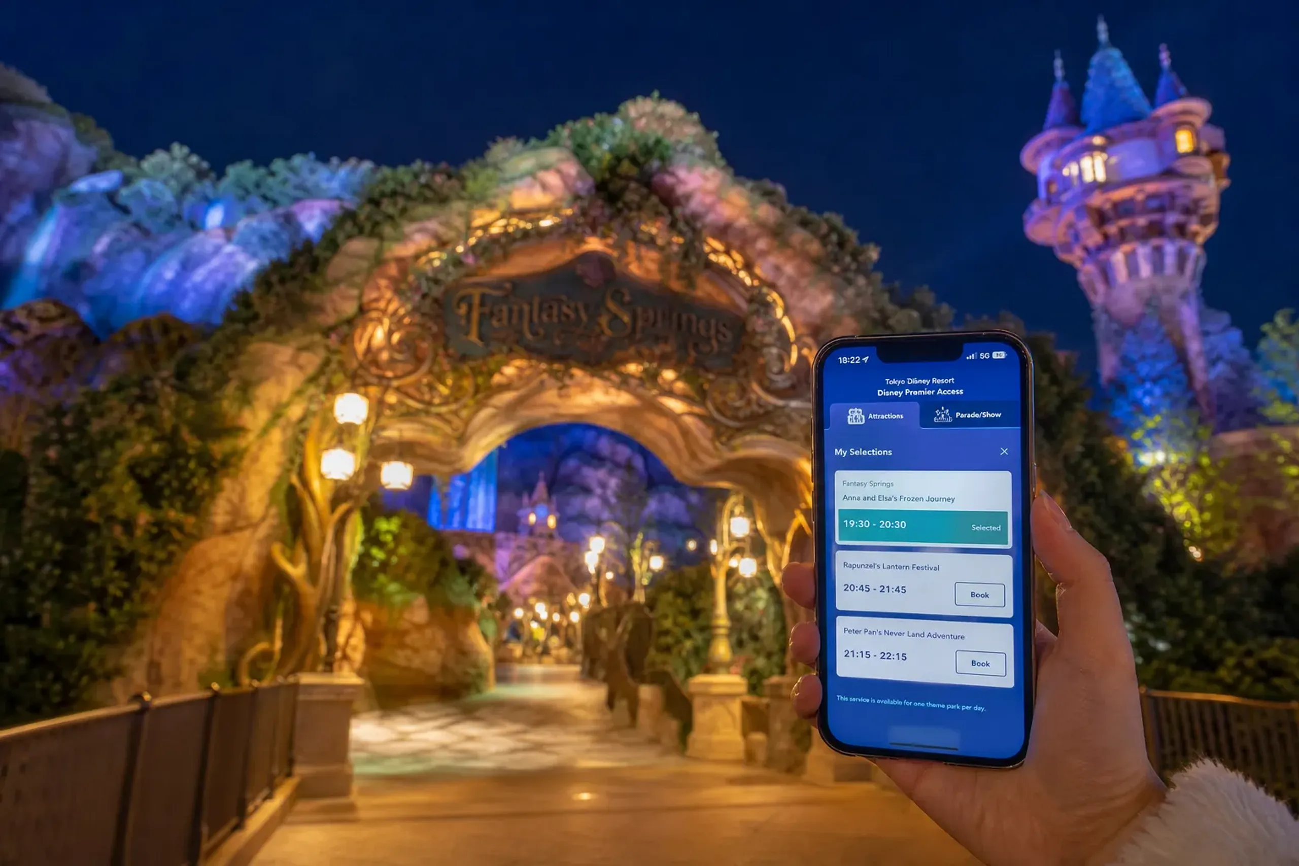 A smartphone displaying the Tokyo Disney Resort app's Disney Premier Access time-slot booking screen, held in front of the softly glowing storybook archway entrance to Fantasy Springs at Tokyo DisneyS