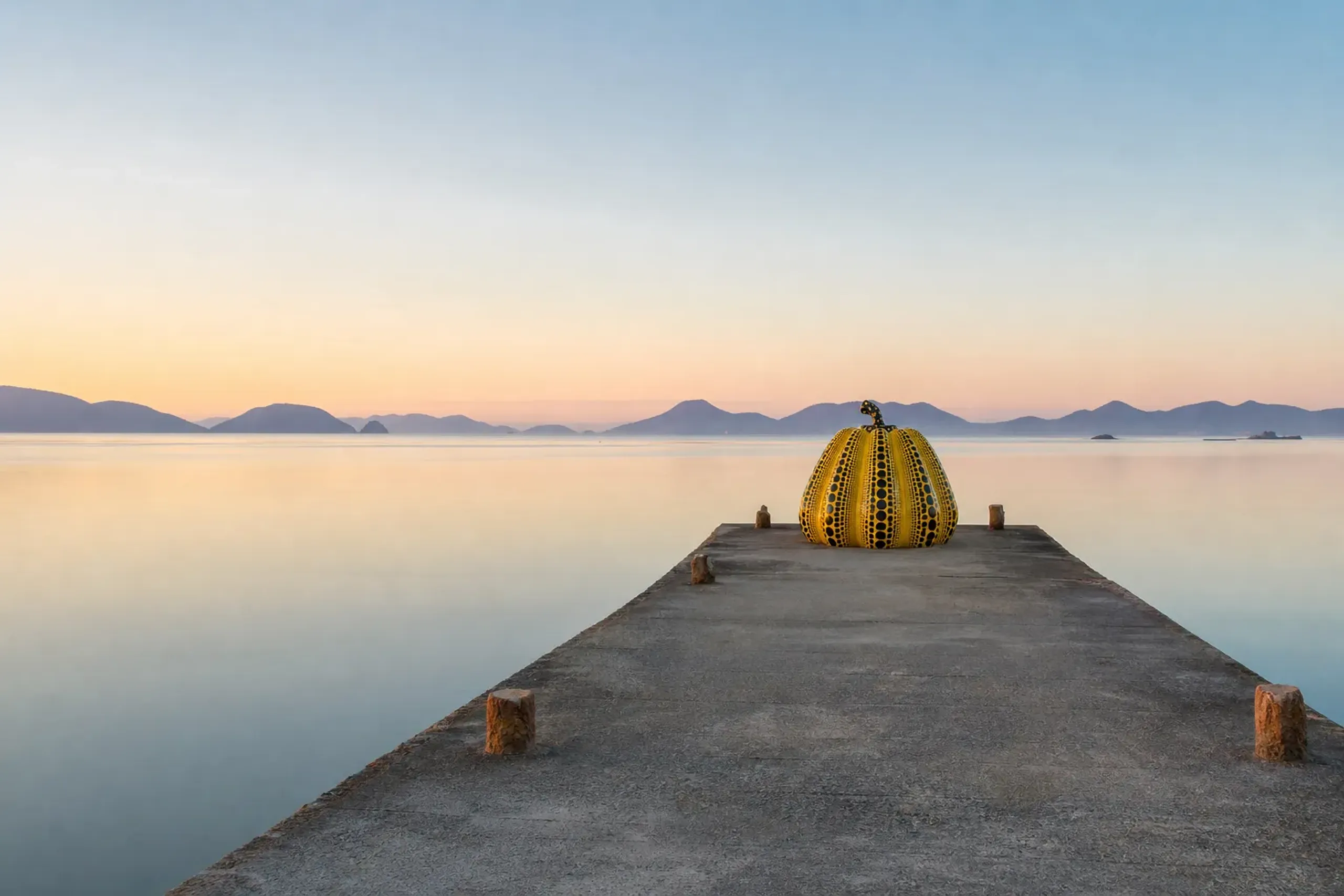 Large yellow and black spotted sculpture on a wooden pier extending over calm blue water with distant mountains on the horizon.