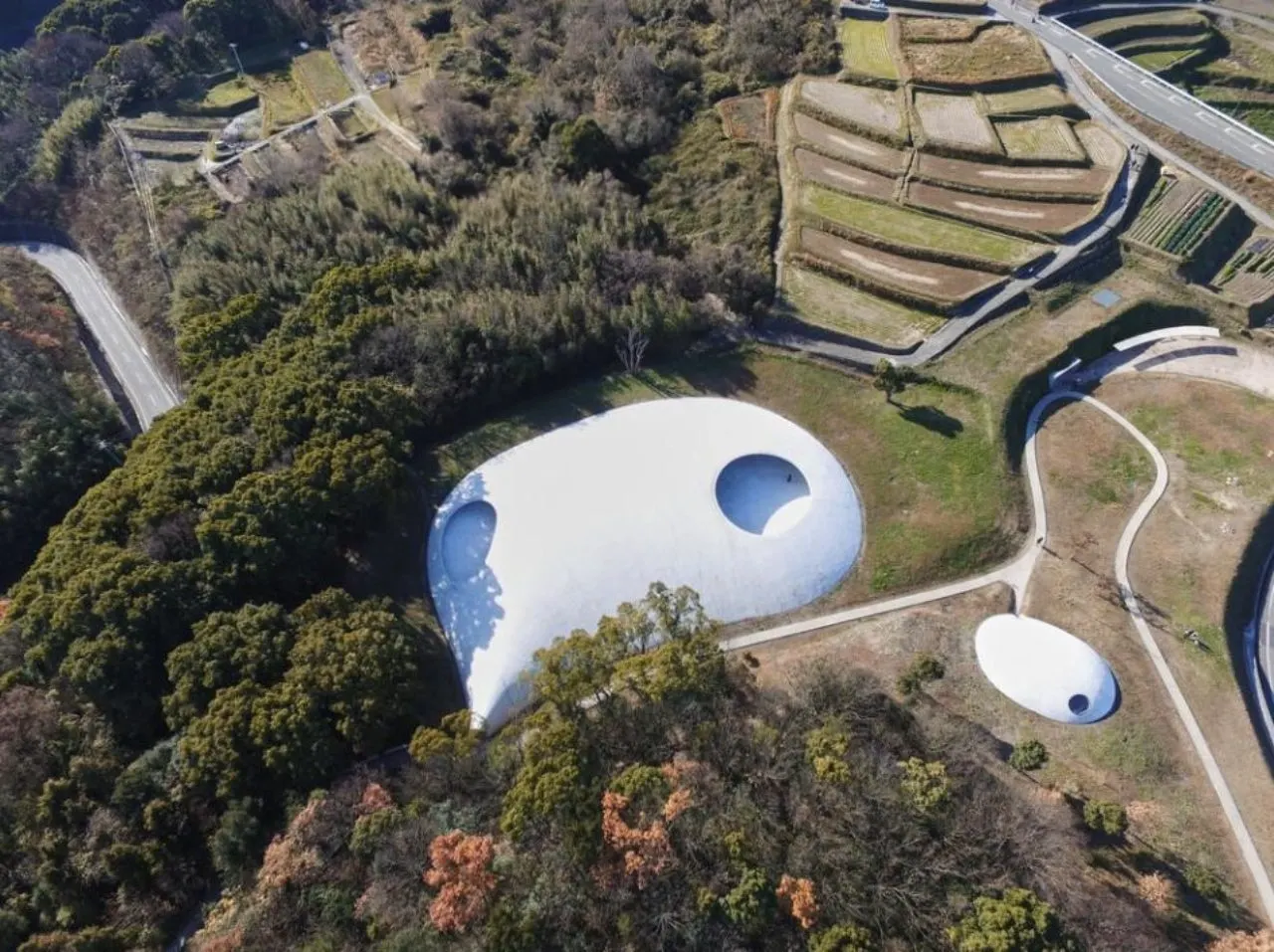 Aerial view of a white dome-shaped building with circular openings, surrounded by trees and curved paths in a park-like landscape
