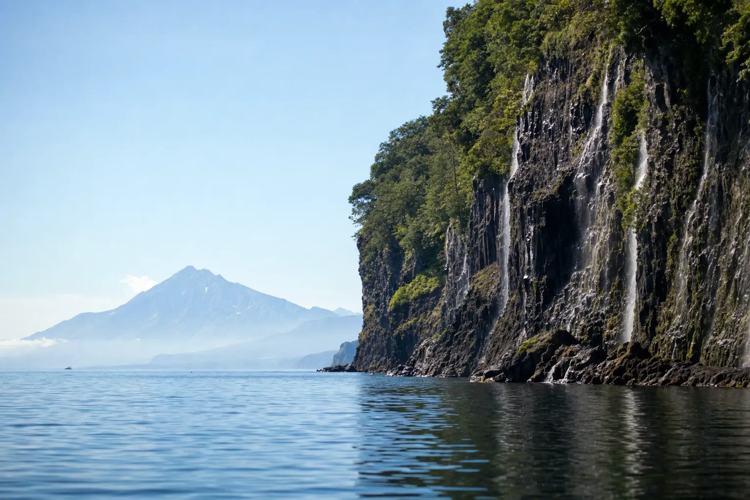the towering coastal rock cliffs of the Shiretoko Peninsula seen from the water, with Mount Rausu rising in the distance under summer light