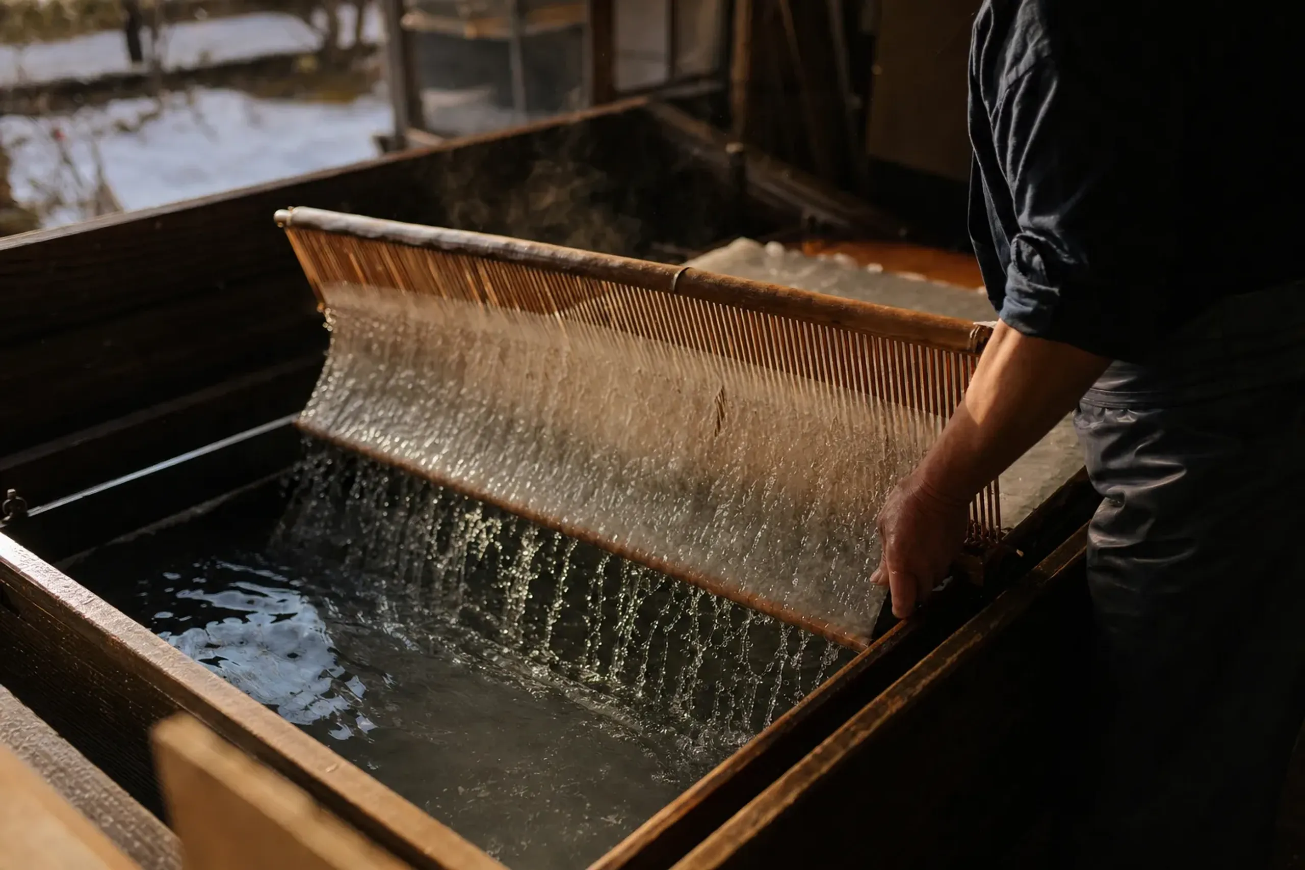 a washi papermaker's bamboo forming screen lifting a sheet of pulp from cold water, caught mid-motion in a winter workshop