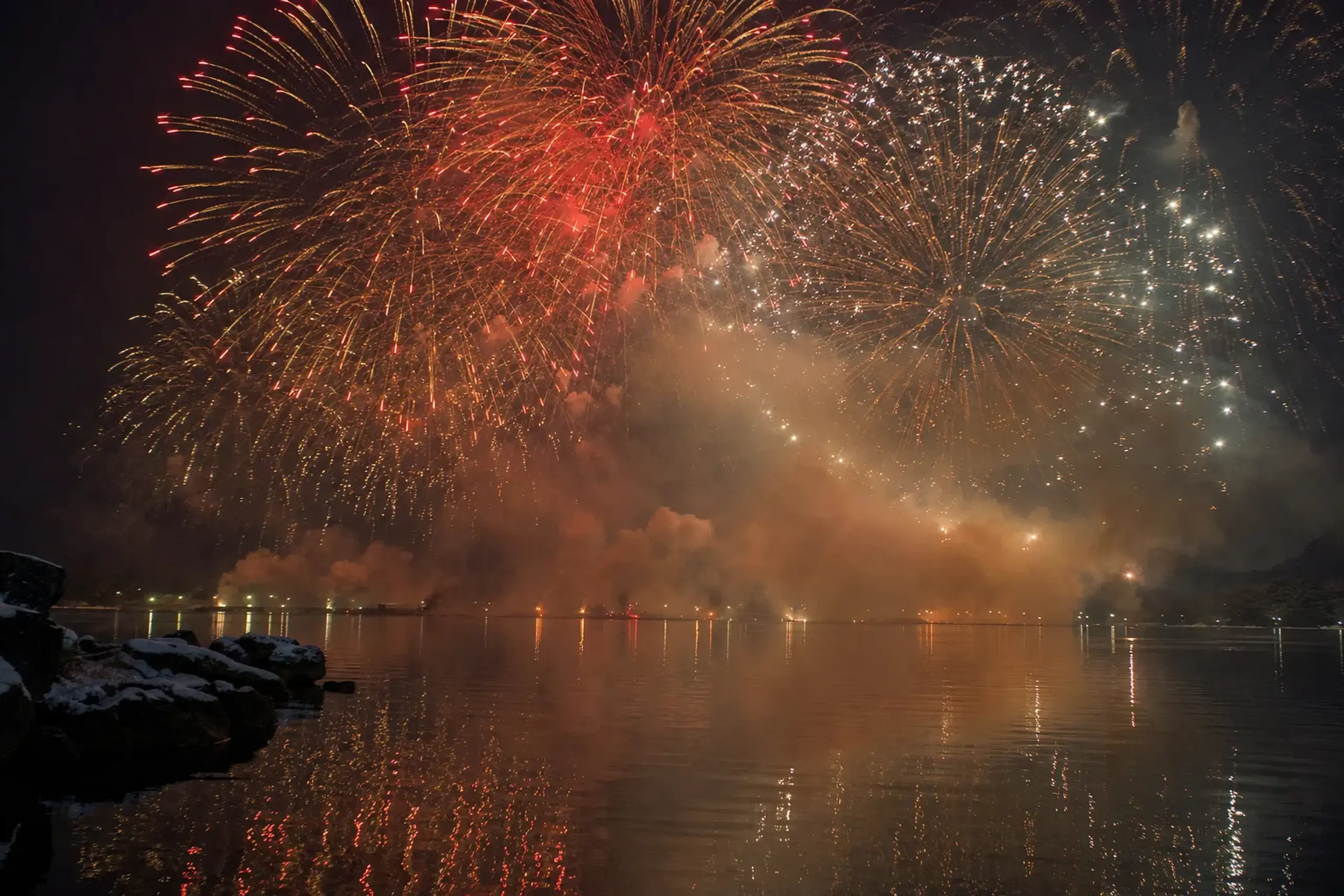 Dense layered fireworks bursts filling the night sky over the ink-dark surface of Lake Suwa in sustained overlapping waves