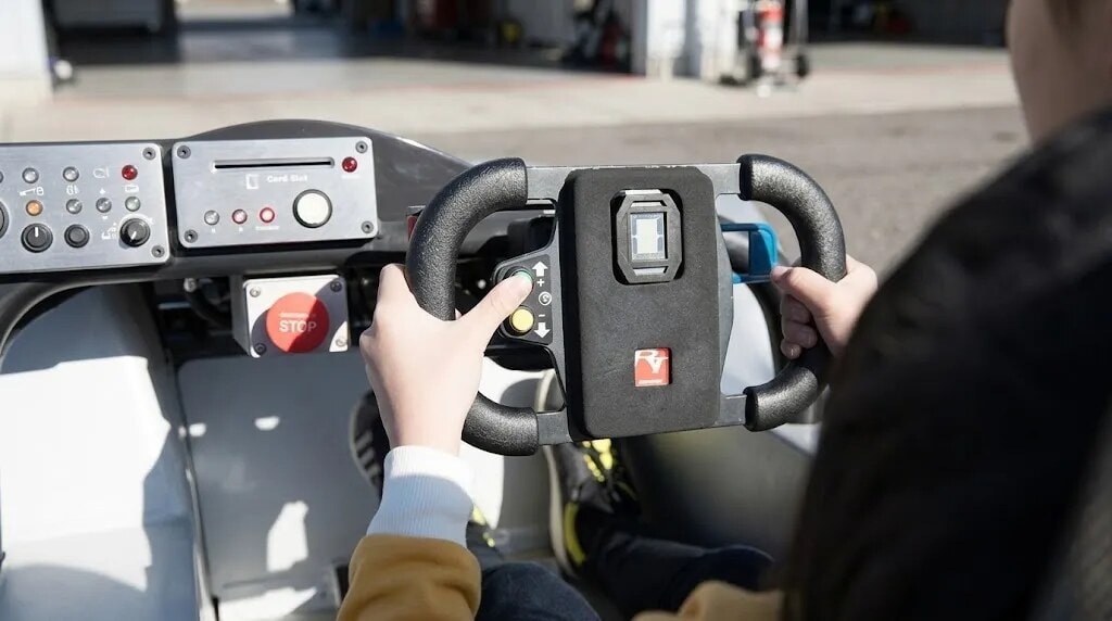 Person's hands on a large steering wheel inside a vehicle cockpit, with a red Stop button and control panel nearby.