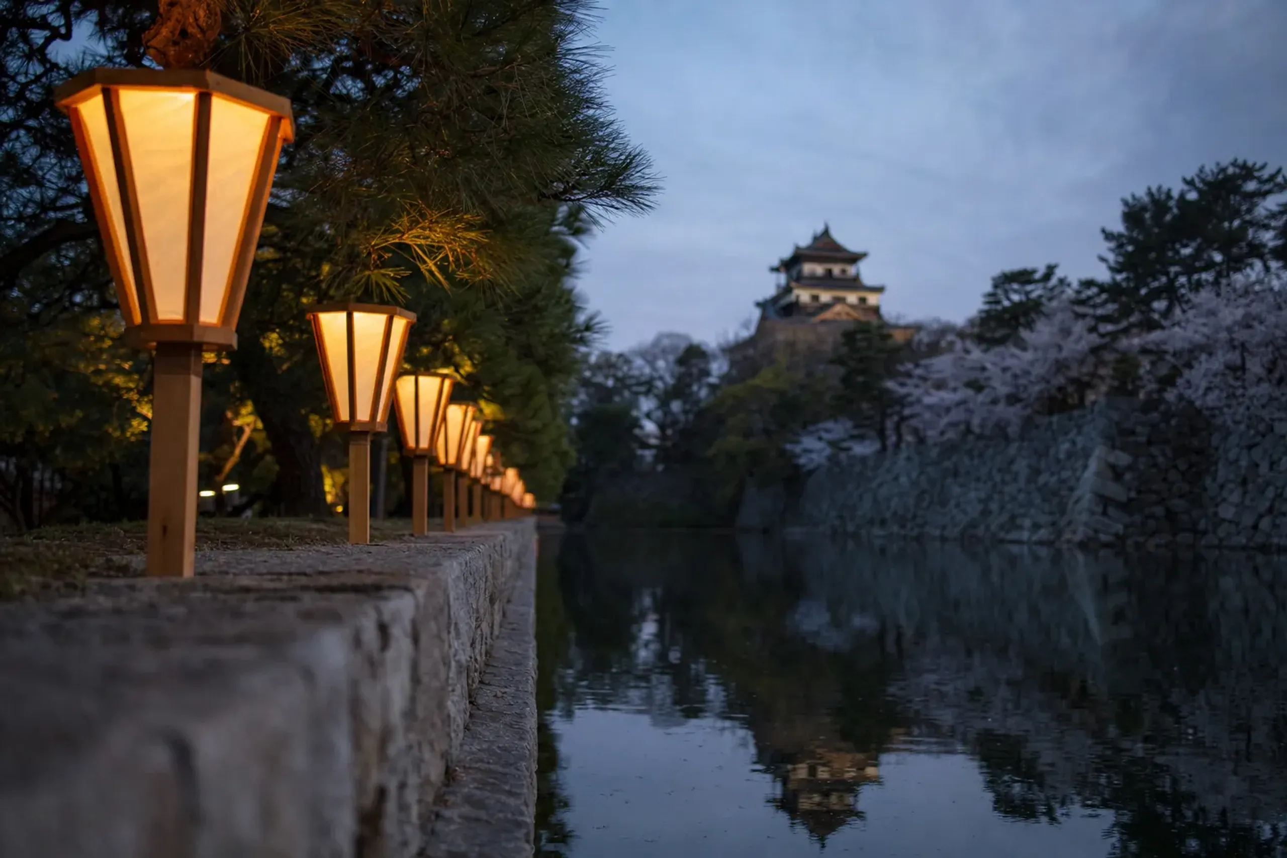a row of glowing paper bonbori lanterns lining the calm castle moat's edge at Takada Castle Site Park at dusk, their soft light reflected on the water