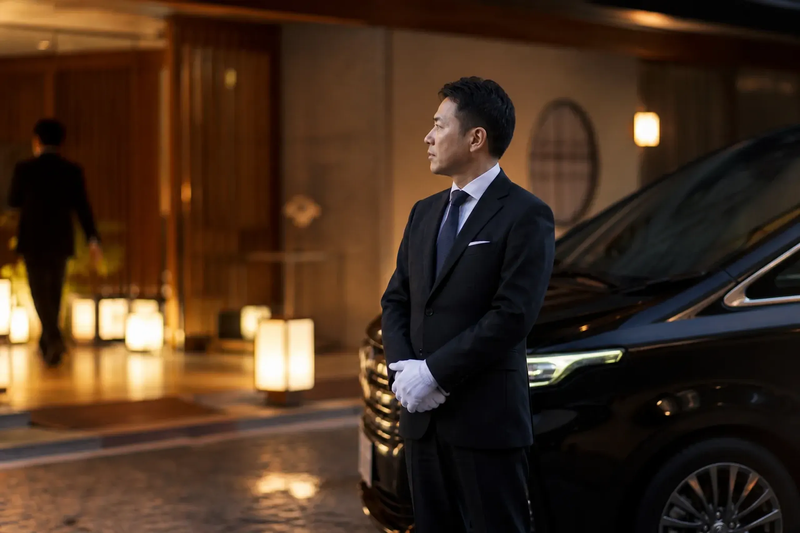 Chauffeur holding a name sign in a Tokyo airport arrival lobby beside a black luxury minivan