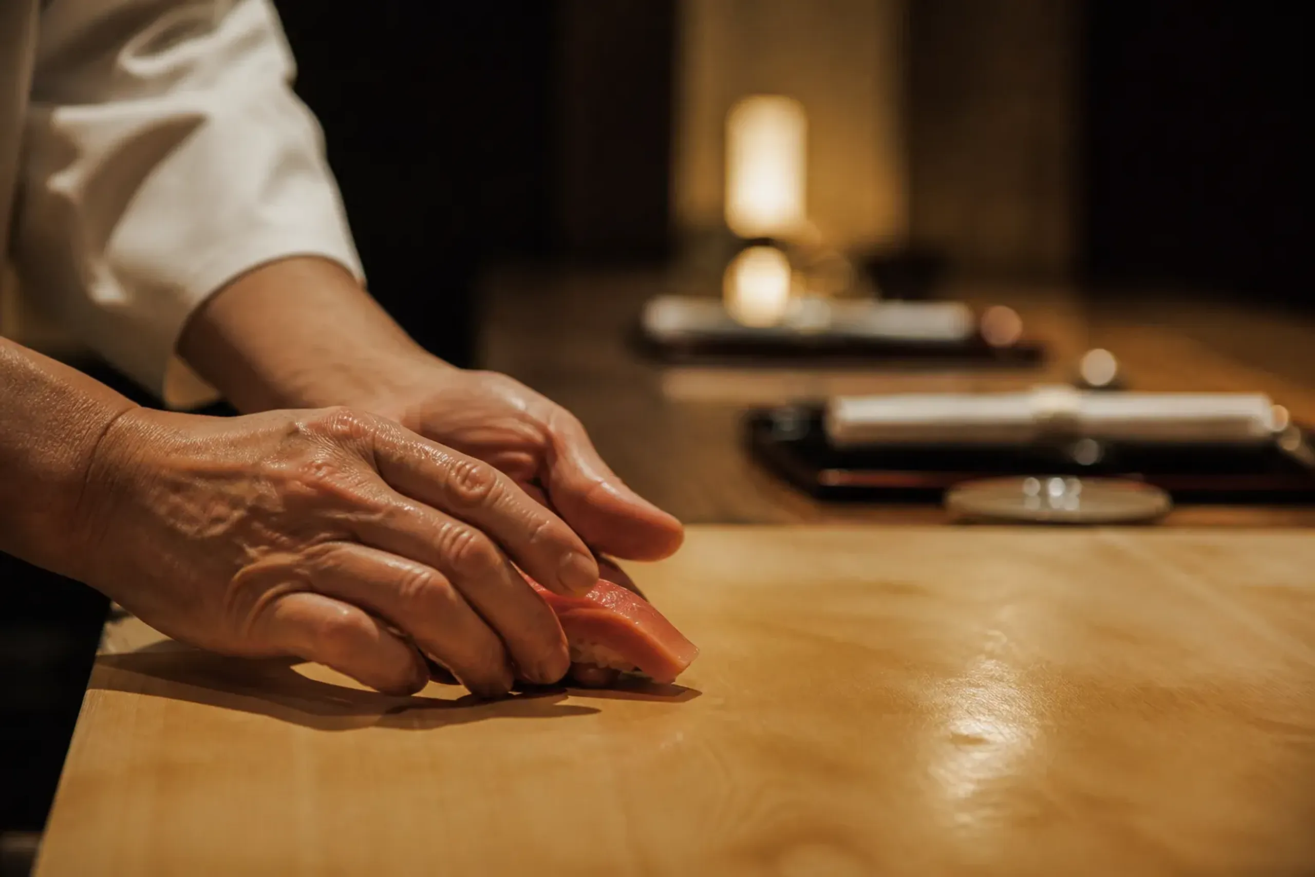 Chef’s hands shaping sushi at a cypress-wood counter with two place settings in warm light