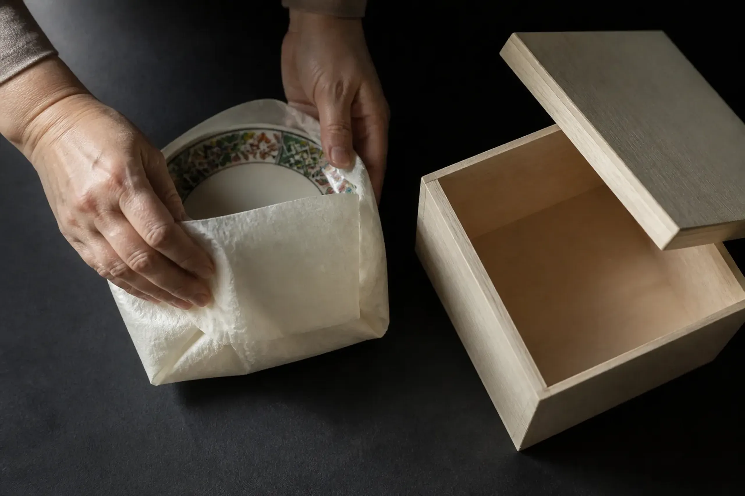 a finished Kutani ware porcelain bowl with polychrome overglaze enamels being wrapped in protective paper and nestled into a wooden paulownia presentation box for shipping