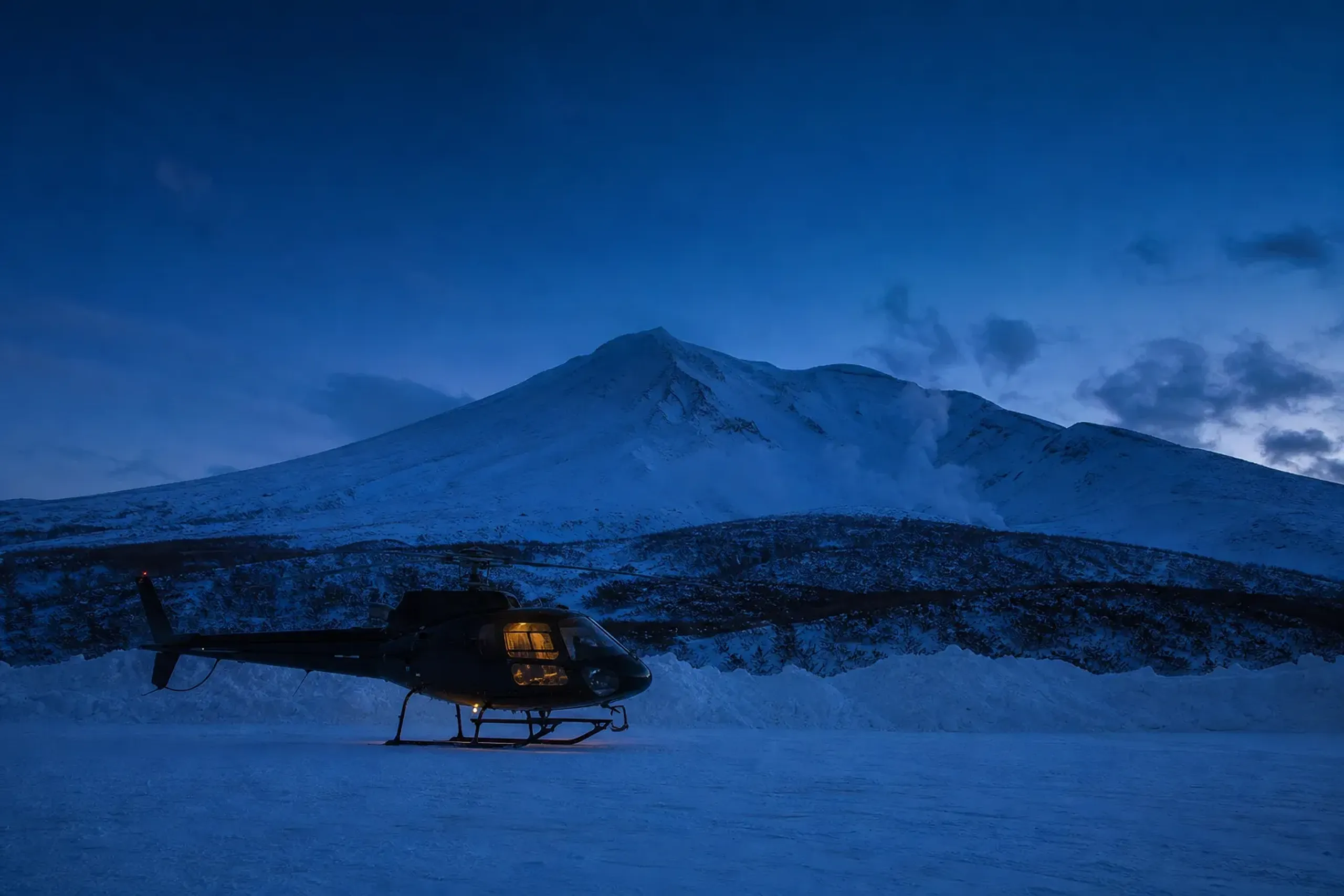 A private helicopter parked on a snow-cleared pad below the peak of Mount Asahidake at dusk in the Daisetsuzan volcanic group, Hokkaido