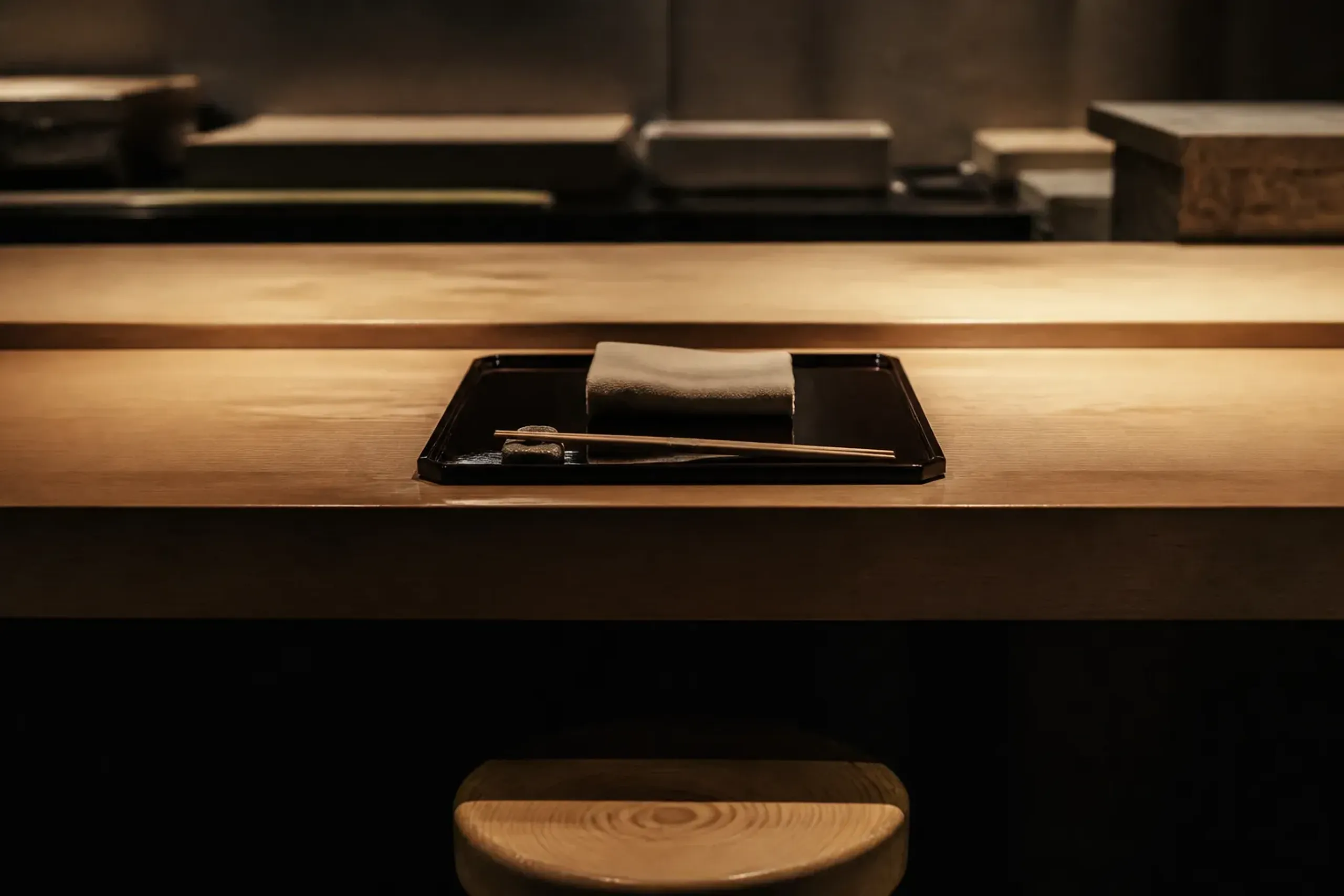 A single low wooden counter seat set for one at a chef-led kappo restaurant in Kyoto, with neatly arranged chopsticks on a ceramic rest and a lone place setting under warm light