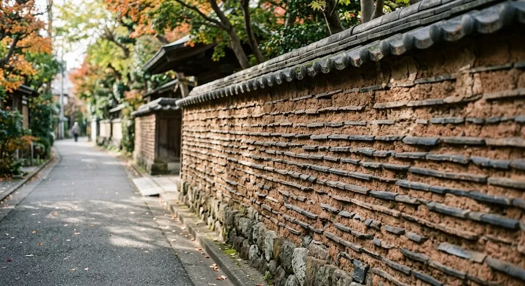 A historic, textured mud and clay wall embedded with rows of dark roof tiles runs alongside a quiet, clean asphalt street under bright daylight in Yanaka, Tokyo.