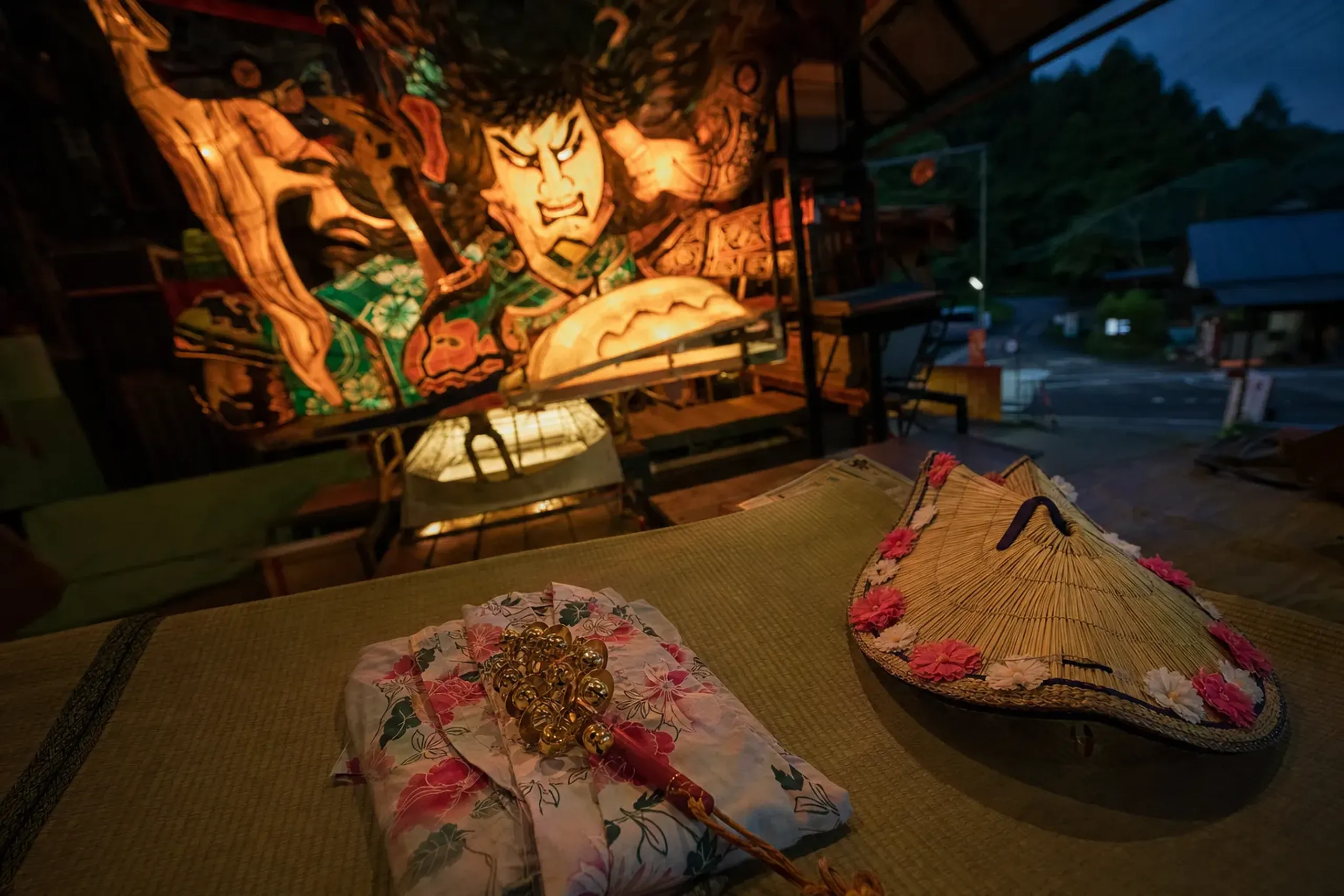A haneto dancer's rental costume with bells and straw hat laid out beside a glowing painted washi Nebuta float