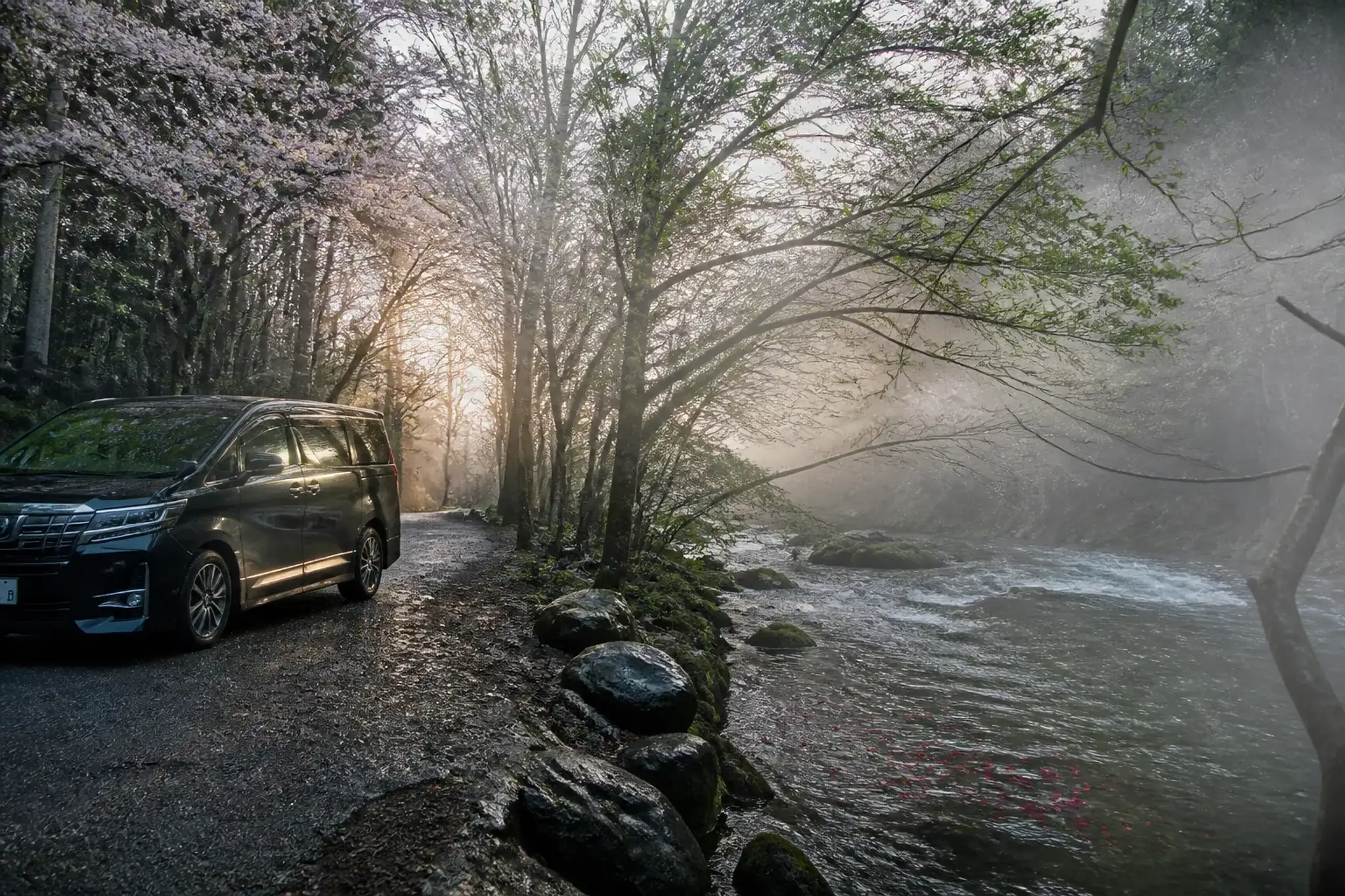A private chauffeur-driven van parked in a quiet pull-off beside Oirase Stream at dawn: empty roadside, pale morning mist, and the stream visible beyond with early red leaves on the current—capturing