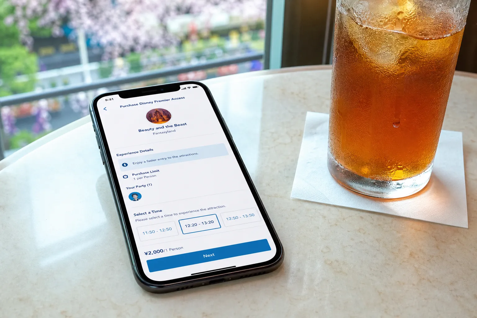 the Tokyo Disney Resort smartphone app open to the Disney Premier Access purchase screen showing a timed attraction return window, resting on a table beside a chilled drink in an air-conditioned Tokyo