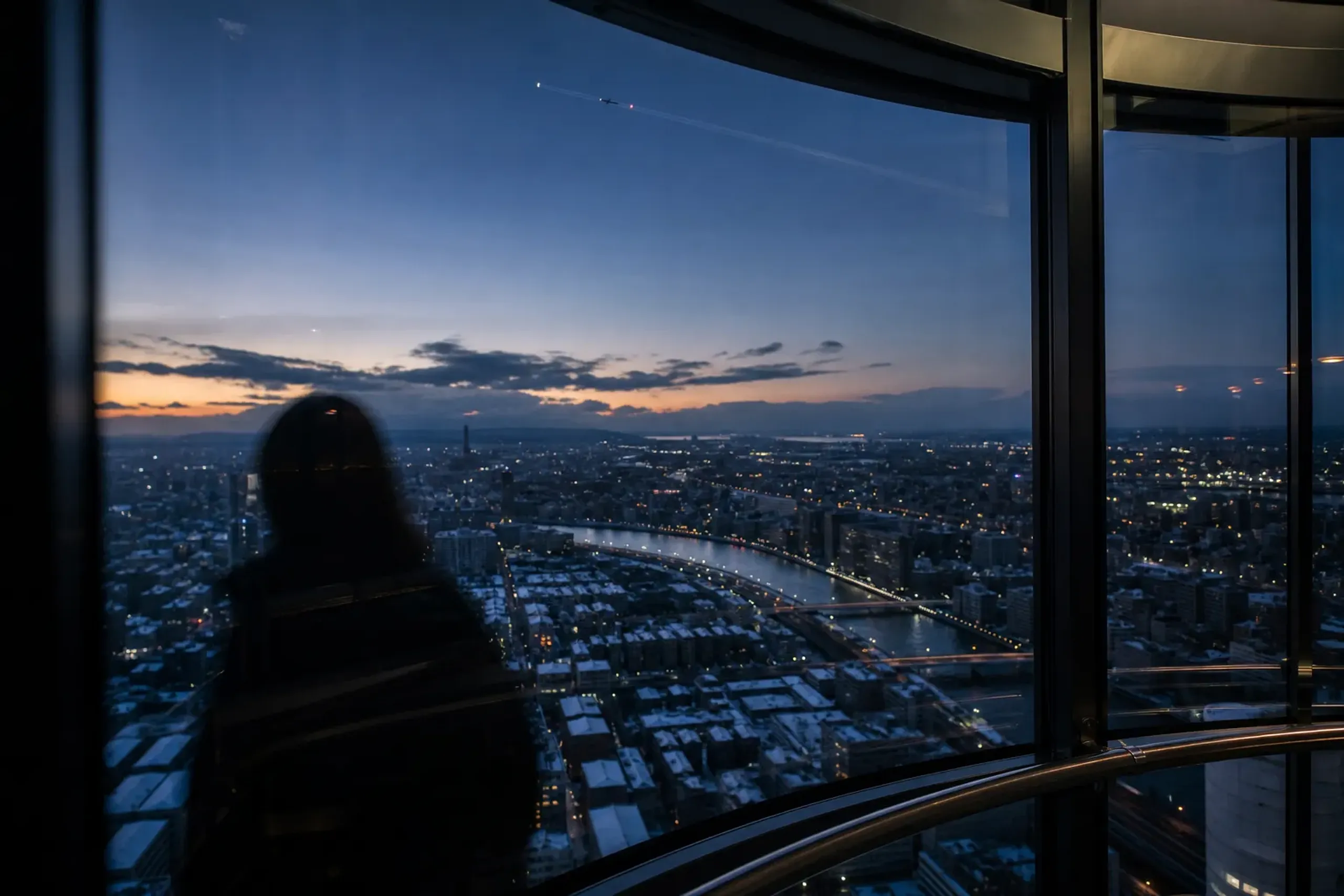 The high-speed glass elevator interior of Tokyo Skytree ascending, with the Sumida River bending far below and aircraft navigation lights blinking in the twilight sky toward Haneda