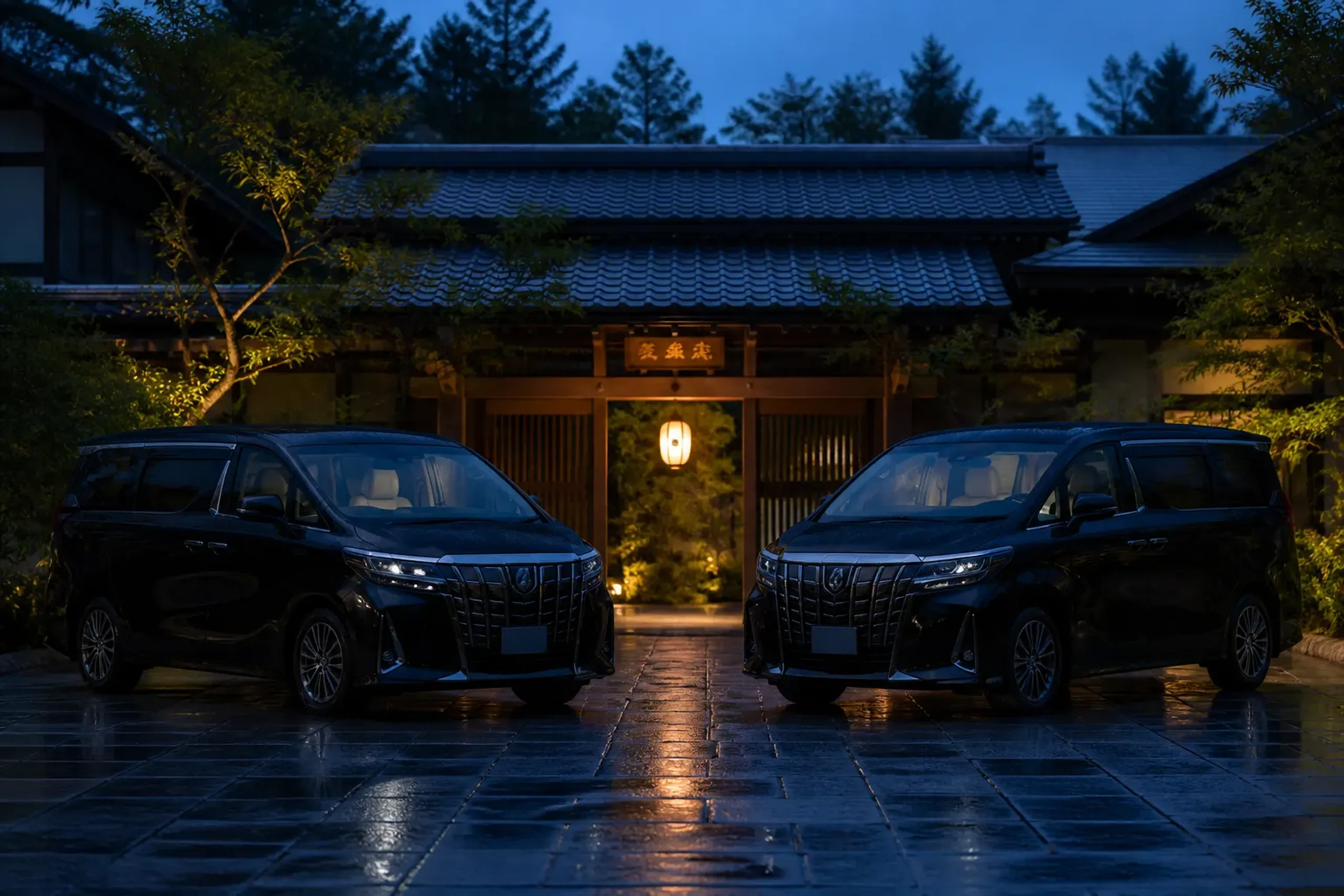 Two matching black luxury minivans parked side by side on a stone-paved driveway outside a traditional Japanese ryokan gate at dusk