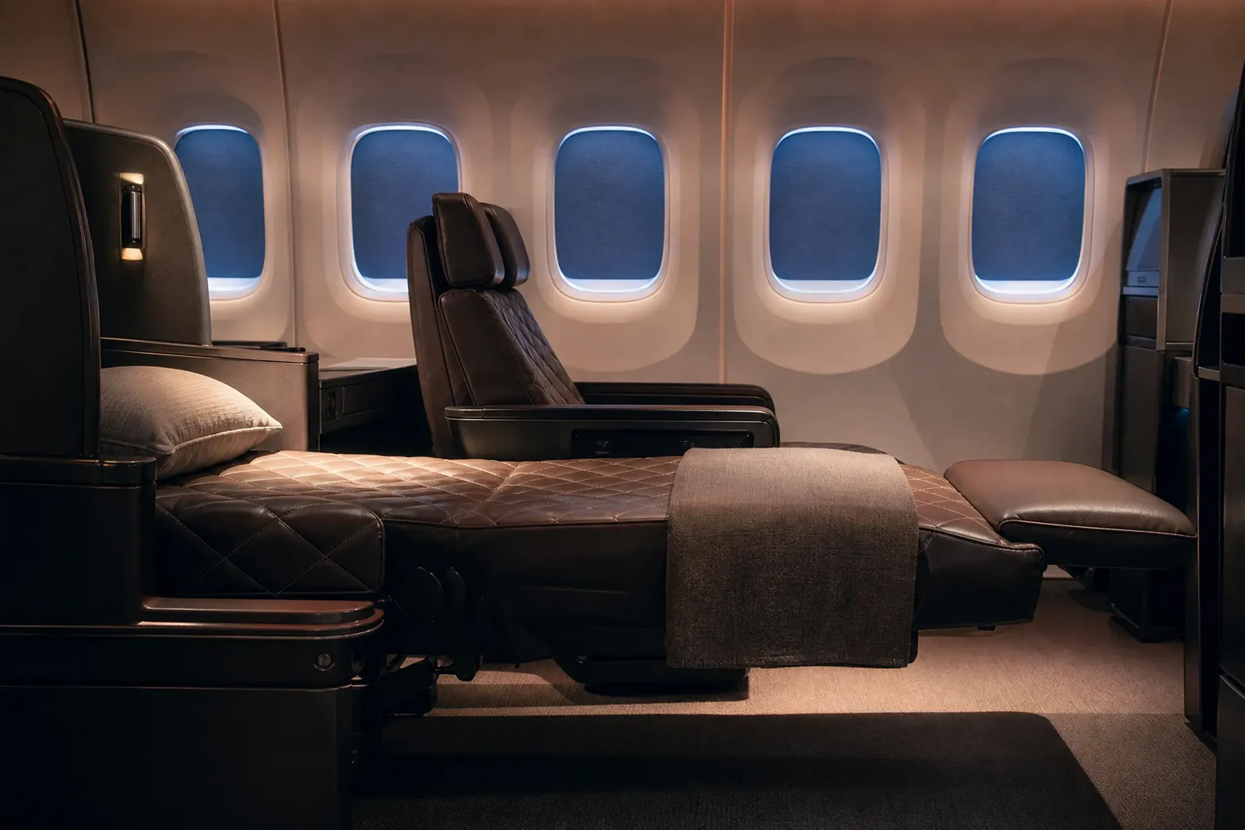 Side view of two dark brown leather seats with diamond stitching in an Airbus A340-300. The closest seat is fully reclined into a flat bed position, connecting with a padded footrest. Four cabin windows with blue shades drawn down are visible in the background.