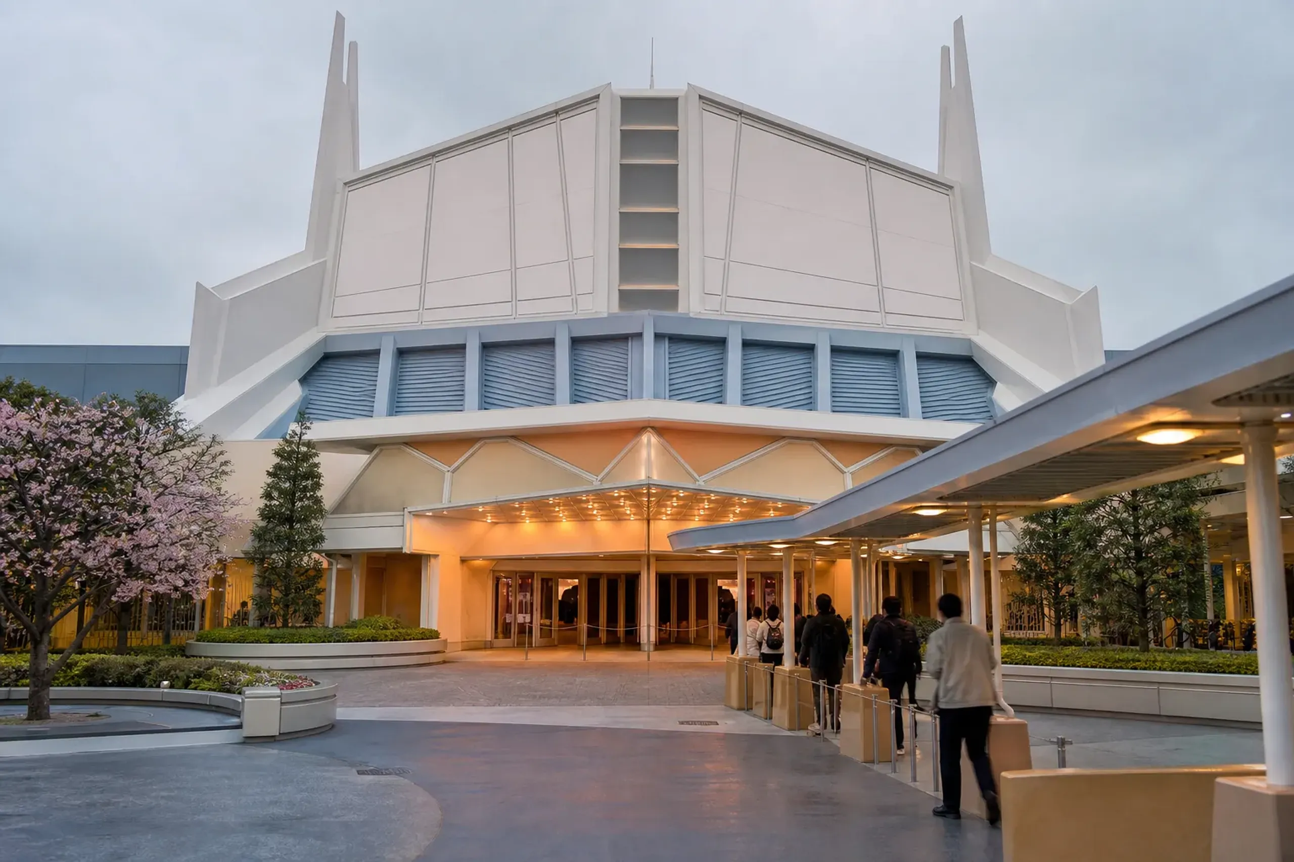The exterior facade and entrance marquee of the Showbase indoor theatre in Tomorrowland at Tokyo Disneyland, with its geometric mid-century-modern architecture and covered queue area under an overcast