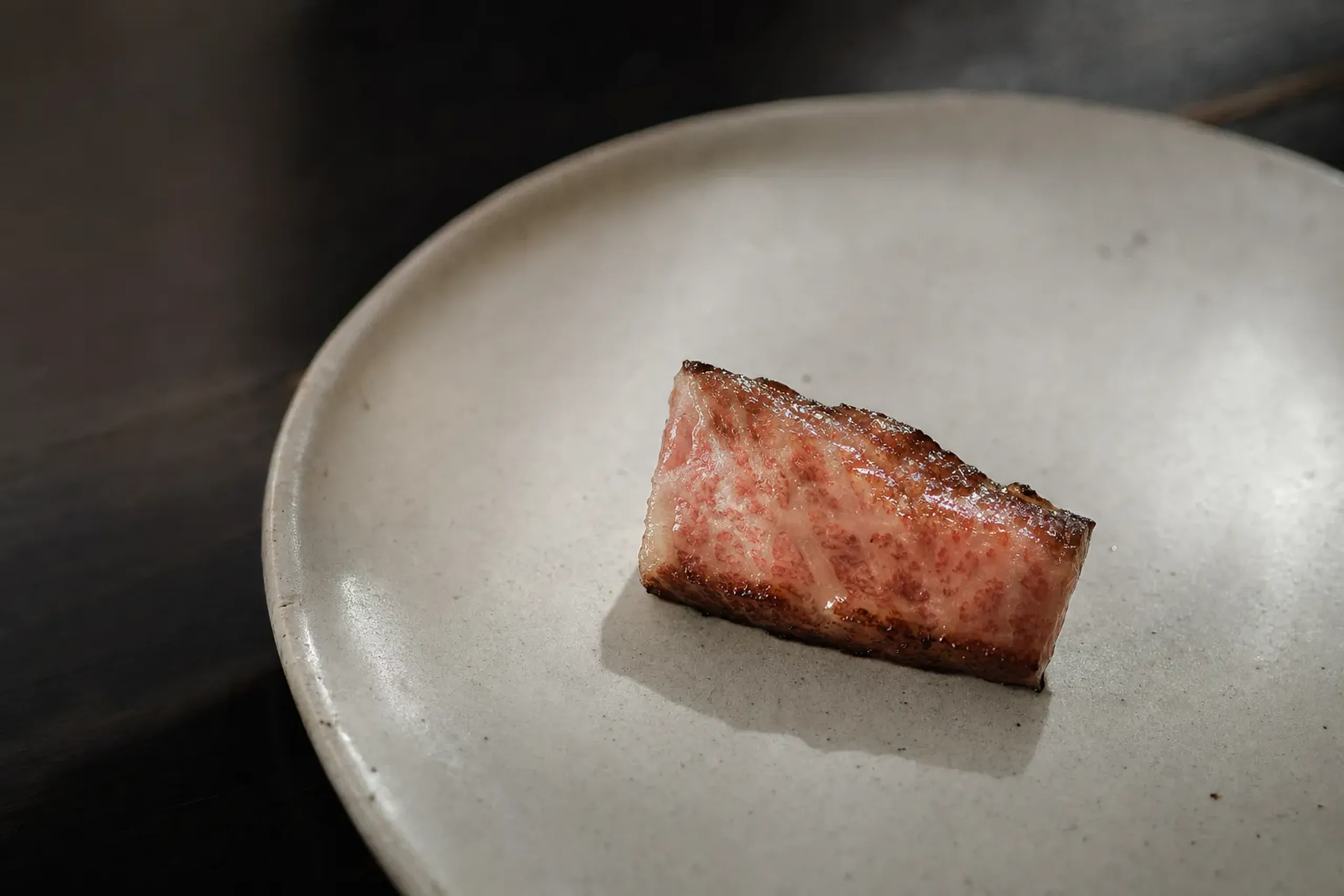 a single seared slice of Tajima-strain Kobe beef with pale glistening fat on a simple ceramic plate