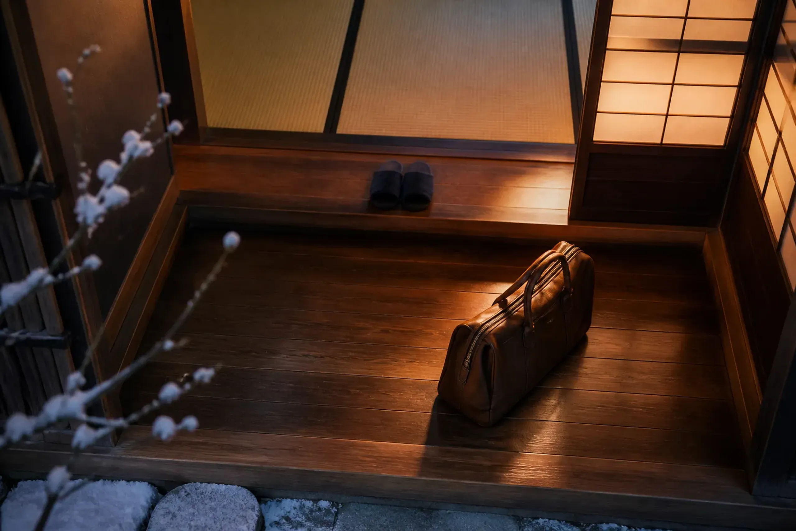 A single leather overnight bag standing alone in a softly lit Kyoto ryokan entryway at dusk, tatami and shoji screens behind it, with a low wooden step and folded slippers set beside it