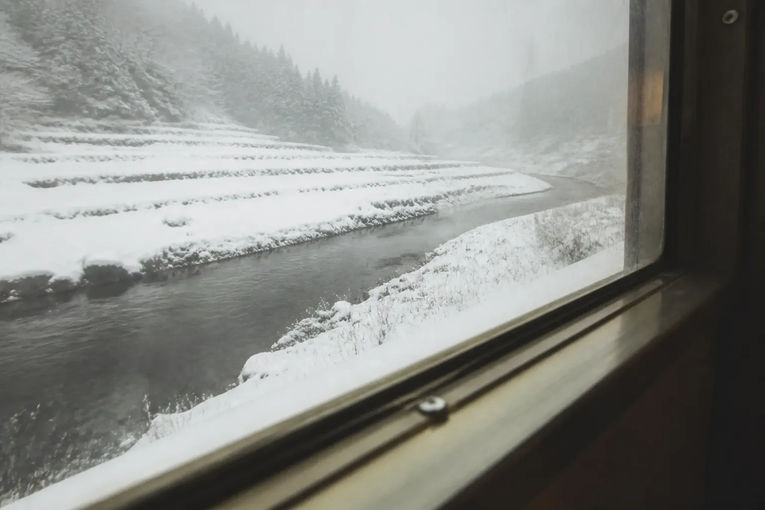 A black river winding through pristine snow country seen from a slow-moving train window, buried rice terraces flattened white to the horizon