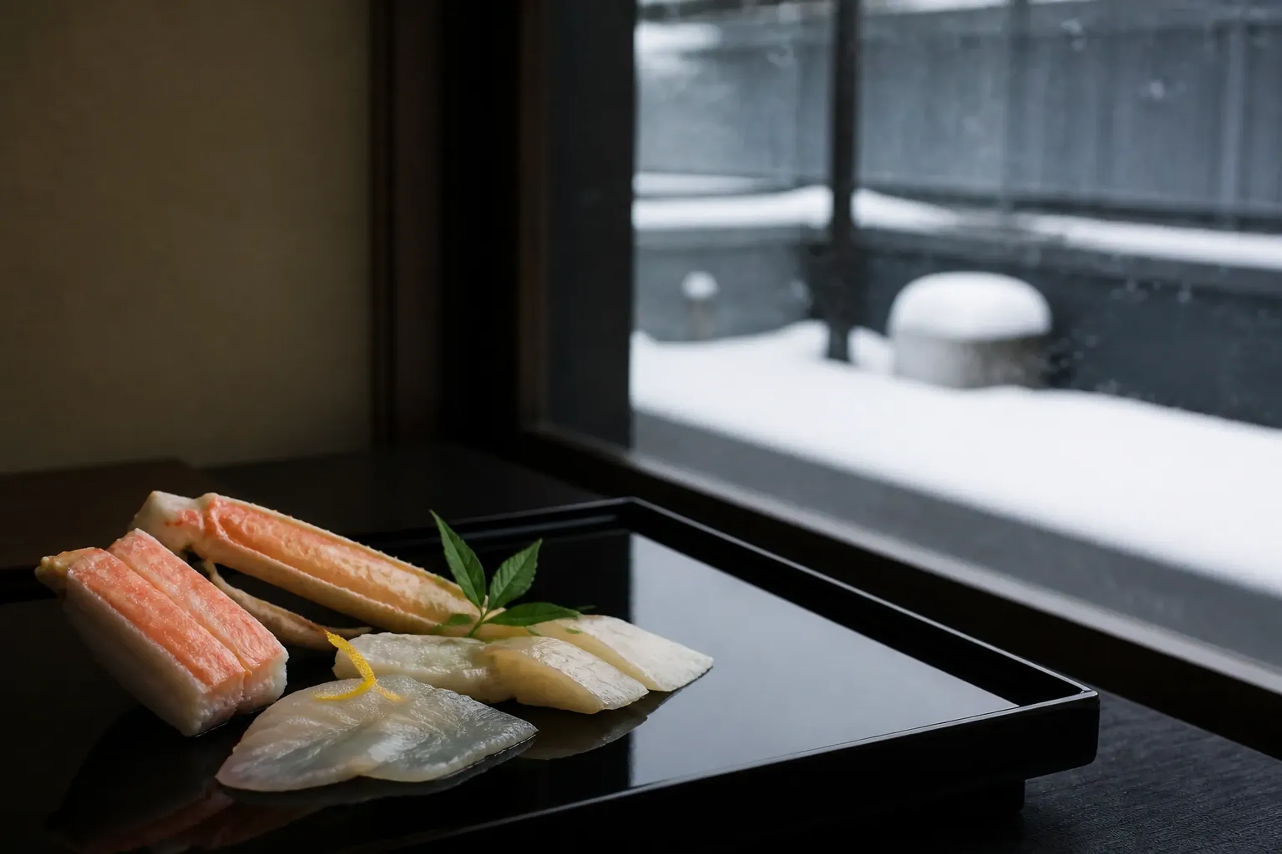 A winter kaiseki dish of cold-water snow crab and firm white fish plated on lacquerware in a snow-region ryokan dining room, snow visible through the window