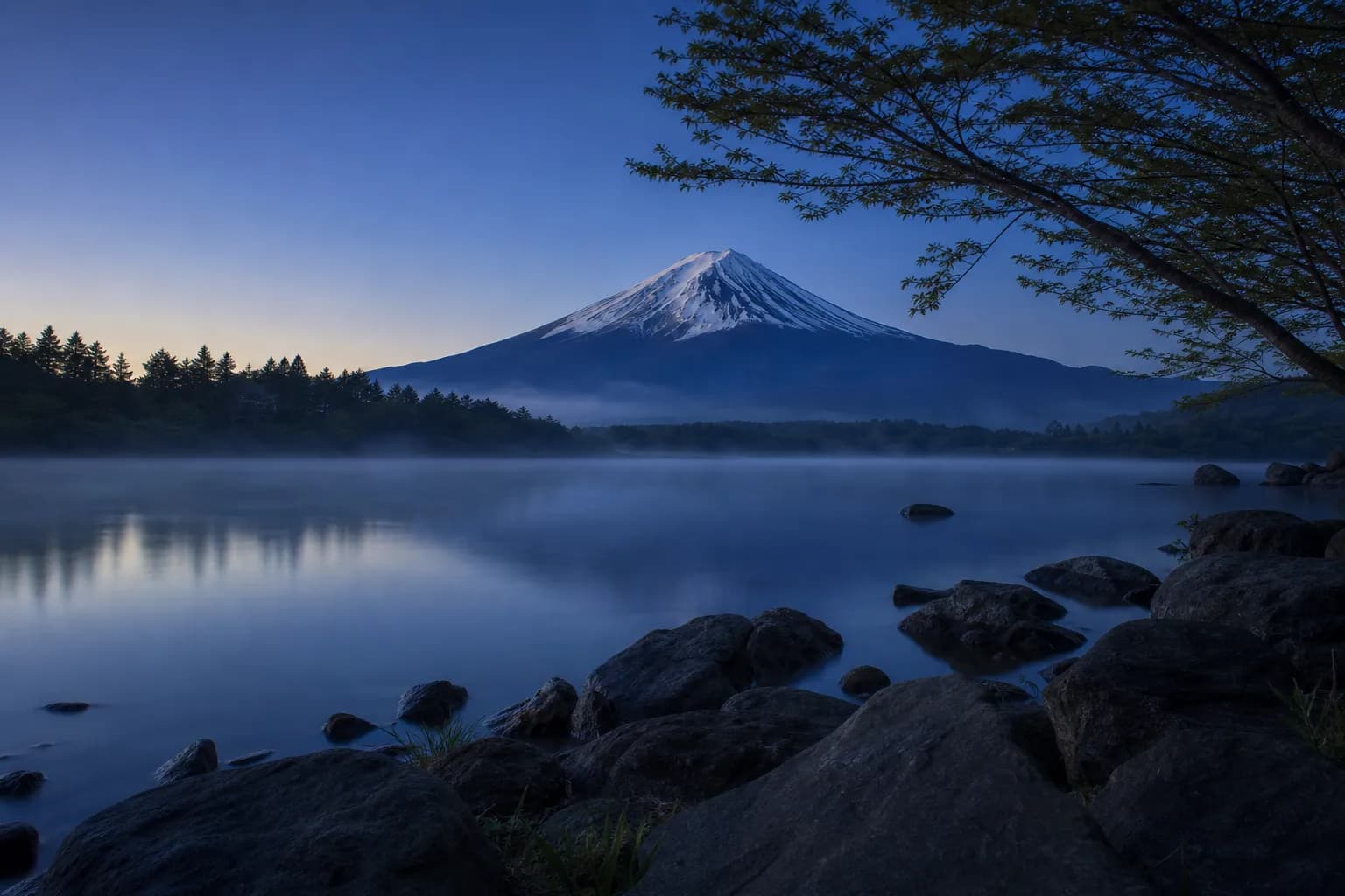 Mount Fuji at sunrise reflected in a calm lake with mist over the forests, snow on the summit
