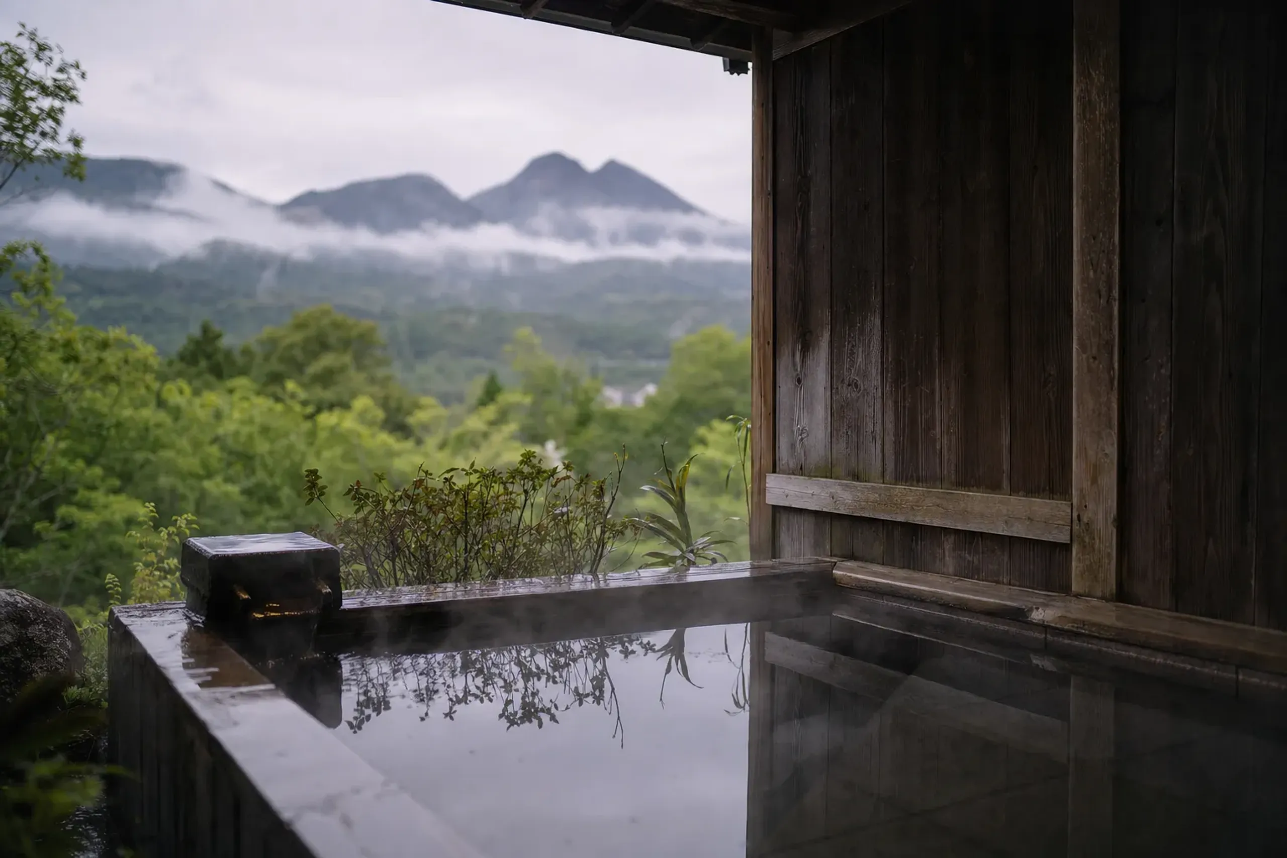 A private outdoor rotenburo bath at a Yufuin ryokan, steam rising from the water, framed by wooden walls and views of Mount Yufu
